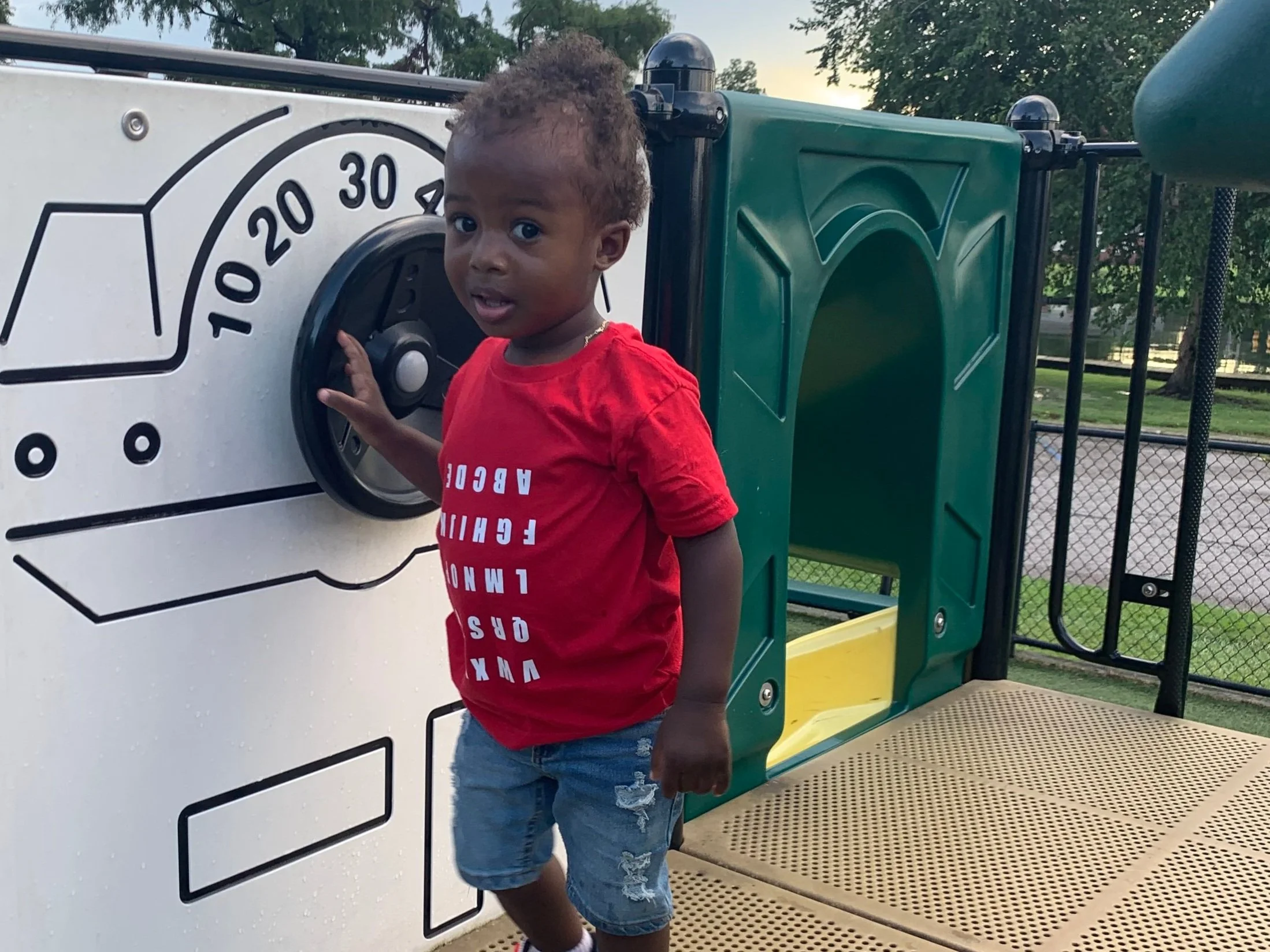 Beautiful brown skinned little boy with curly hair wearing the ABC Tee in red while standing on a playground.