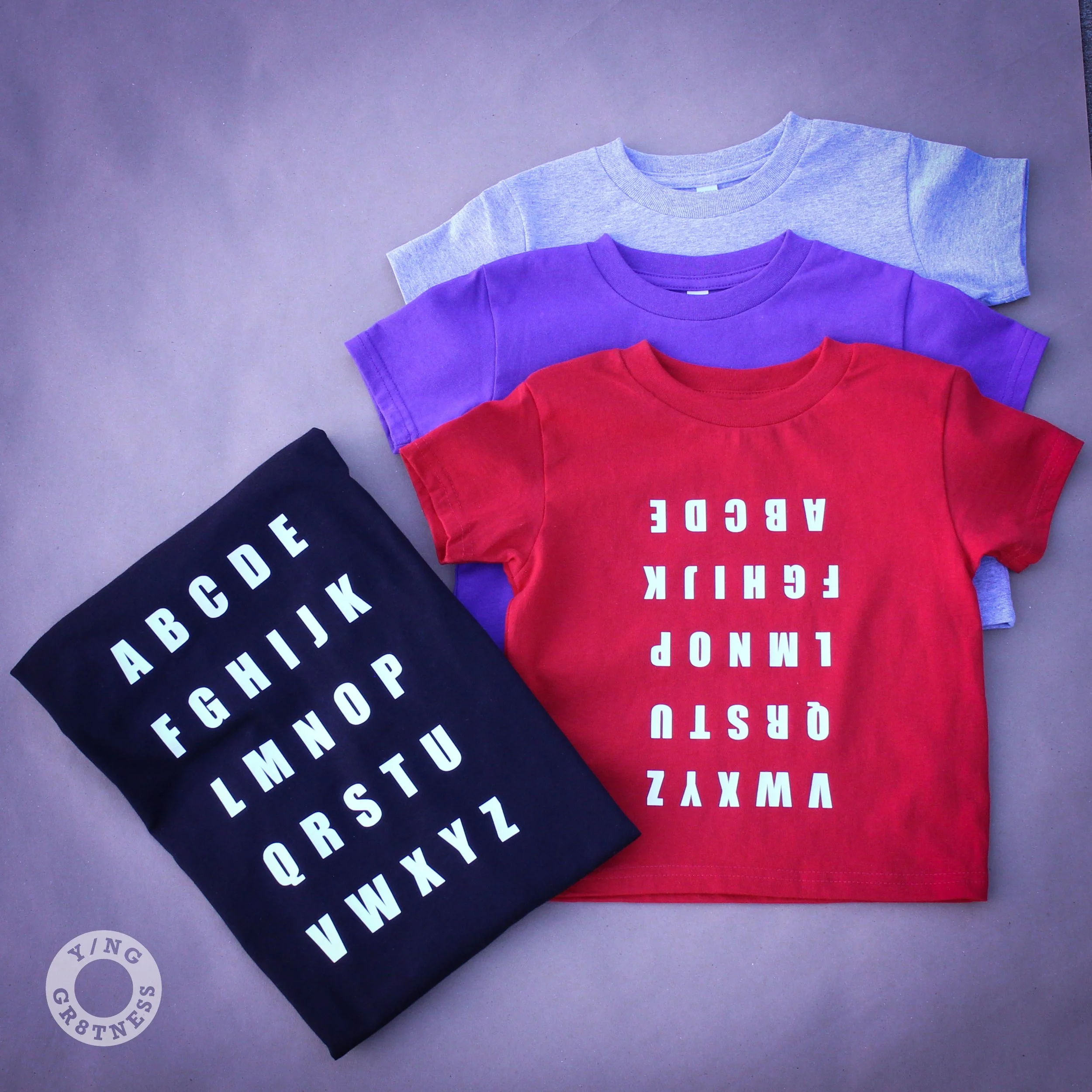A display of the grey, purple and red ABC Tees as well as an adult size Alphabet Graphic Tee.