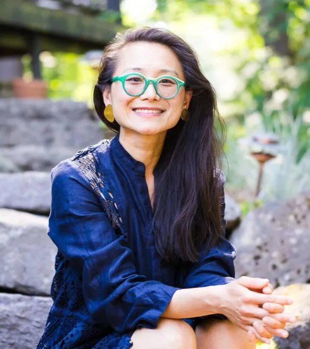 A woman with long dark hair, green glasses, and gold earrings smiling outdoors, sitting on rocks in a garden with green foliage.