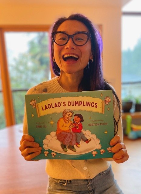 A woman with glasses and long dark hair smiling and holding a children's book titled 'Laolao's Dumplings'. She is wearing a cream-colored sweater and standing in a bright room near a window.