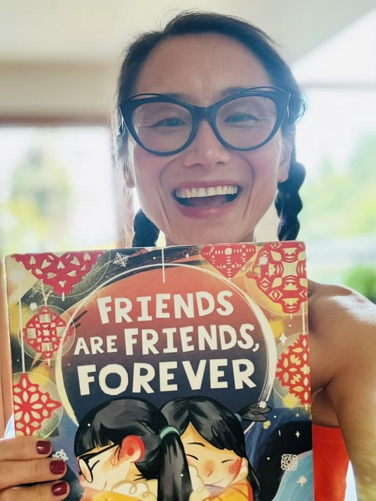 A woman with glasses and braided hair smiling, holding a colorful book titled "Friends Are Friends, Forever" with an illustration of two girls hugging.
