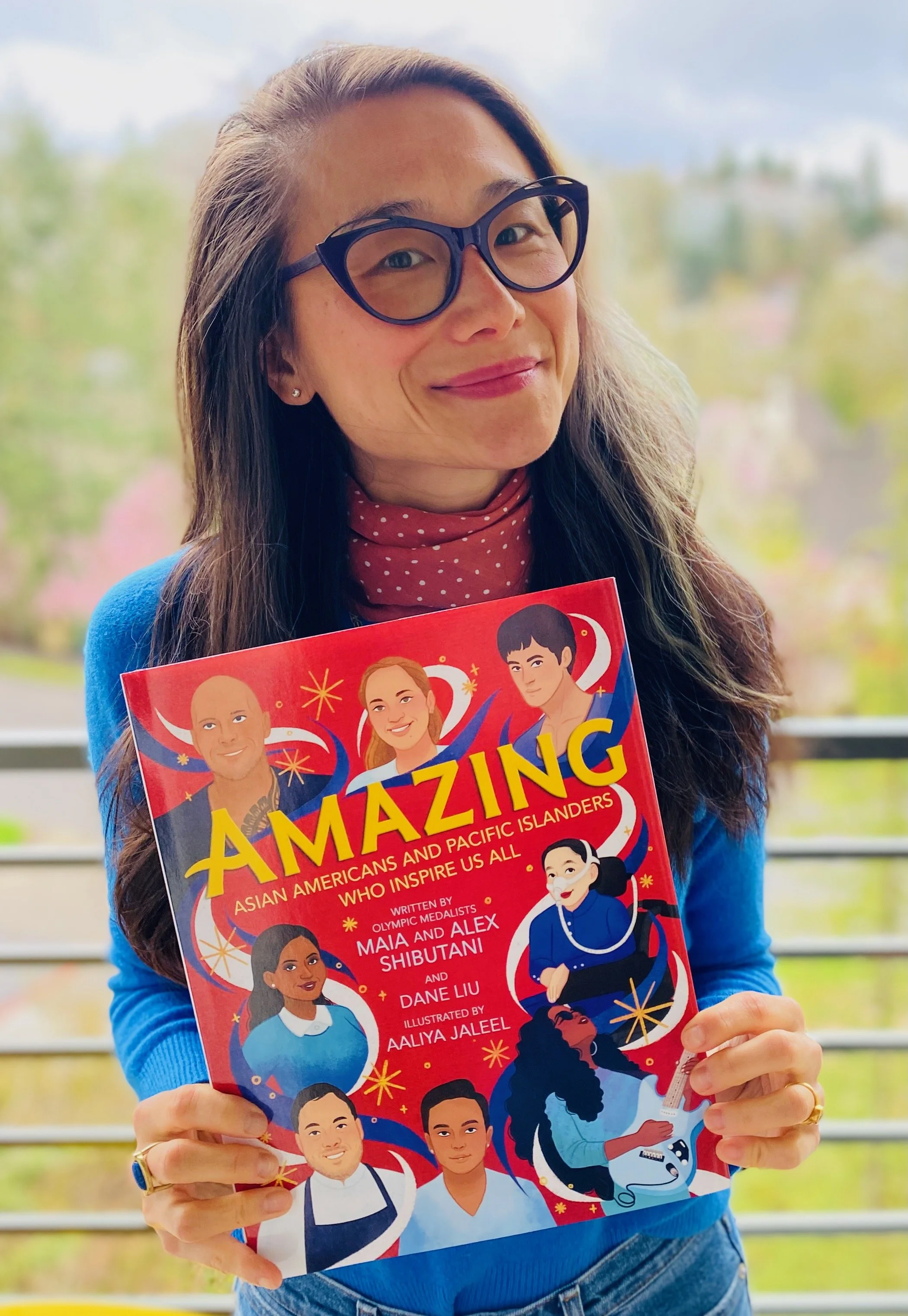A woman wearing glasses and a red polka dot scarf holding a colorful book titled "AMAZING" about Asian Americans and Pacific Islanders, with illustrations of diverse individuals on the cover, standing outdoors with trees and a cloudy sky in the backg