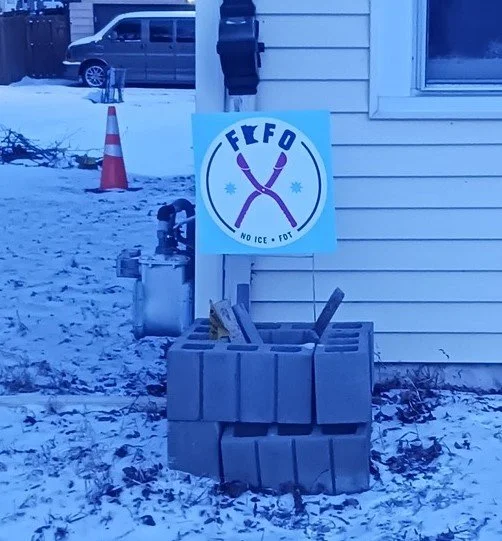 An anti-ICE sign is placed above a stack of cinder blocks at the corner of a yellow house. Snow covers the ground around the house.