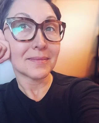 Close-up of a woman with short dark hair, wearing glasses and a black shirt, looking at the camera with a slight smile.