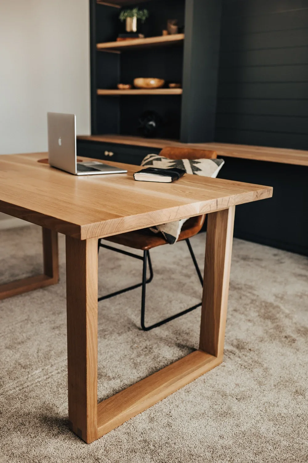 A wooden desk with an open laptop and closed notebook on it. A chair with a patterned blanket draped over it is in front of the desk. In the background, there is a dark blue cabinet with wooden shelves holding decorative items.