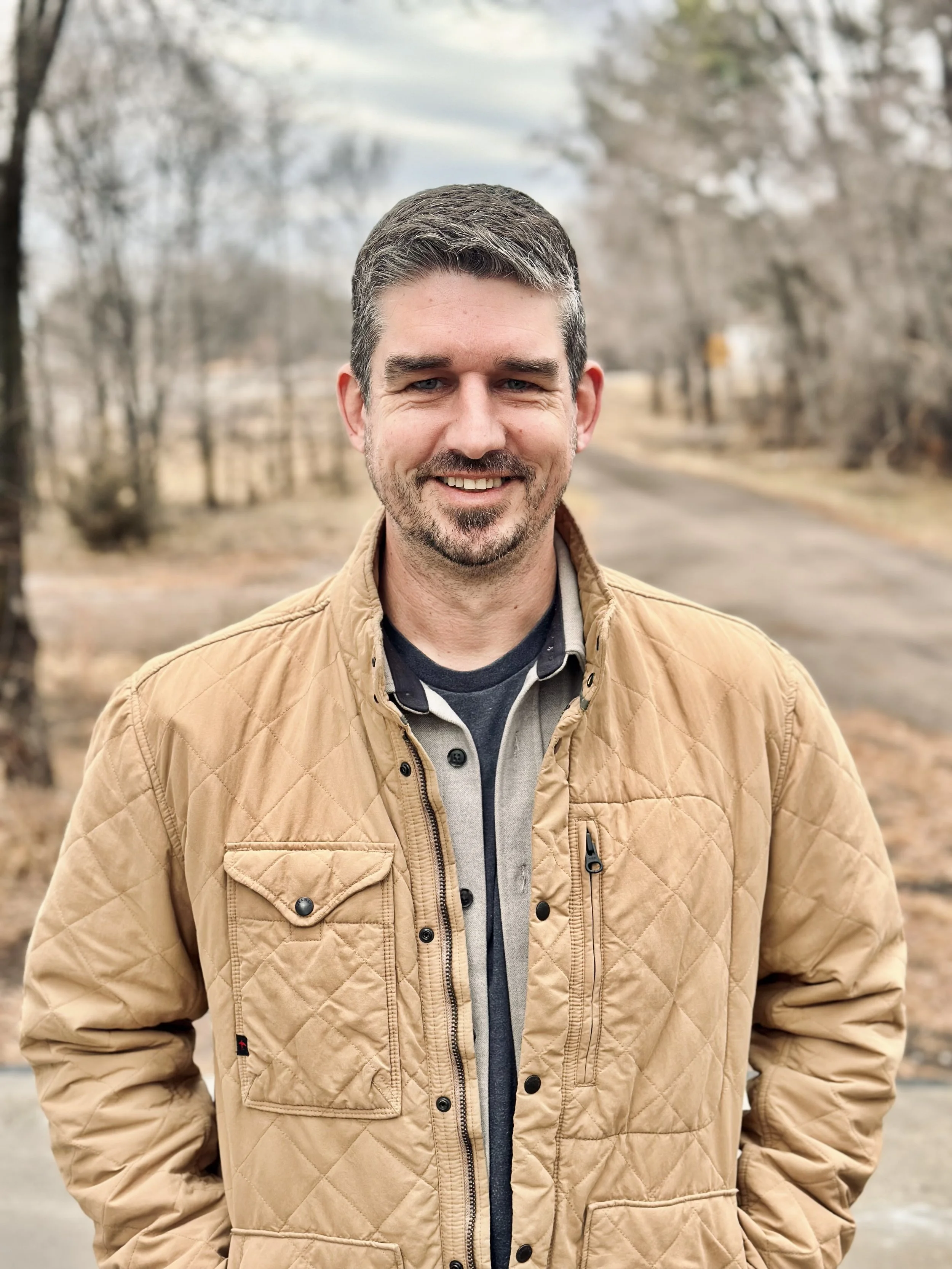 A smiling man in a tan quilted jacket standing outdoors on a leafless tree-lined path during fall or winter.