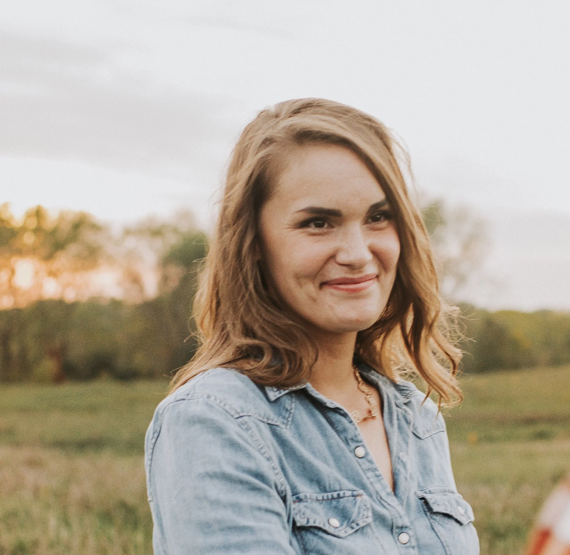 A woman outdoors with a gentle smile, wearing a denim shirt, in a natural setting during sunset.