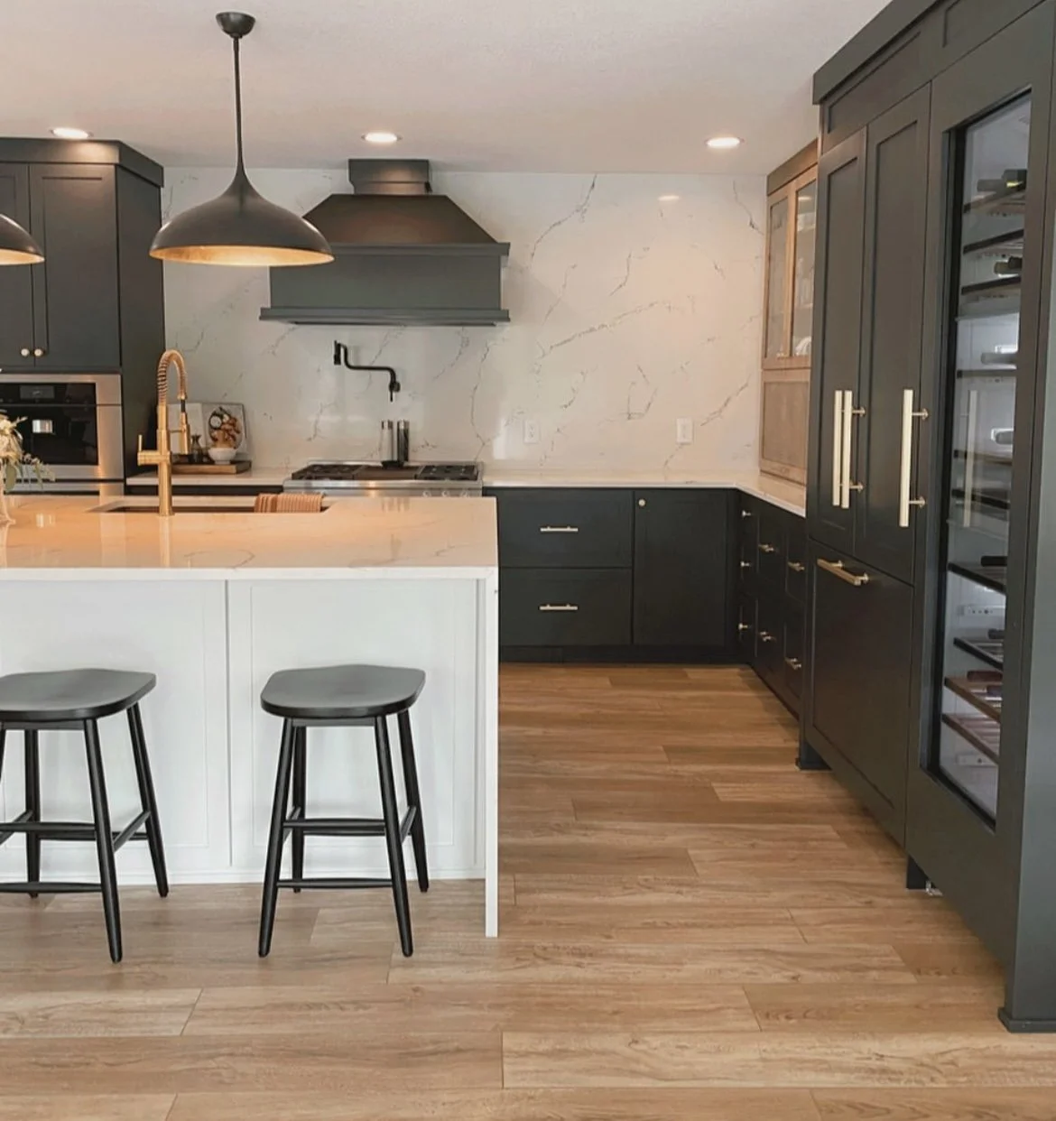 Modern kitchen with black cabinets, wood flooring, white marble backsplash, island with white countertop, black bar stools, stainless steel appliances, and black pendant lights.