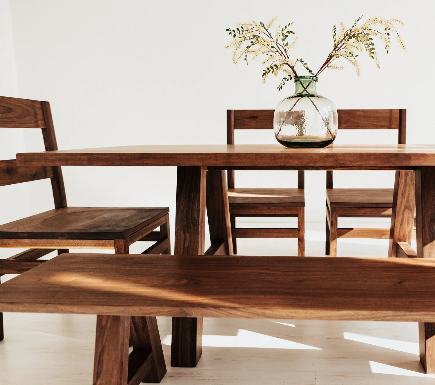 A wooden dining table with a glass vase containing branches and greenery, set against a plain white wall.
