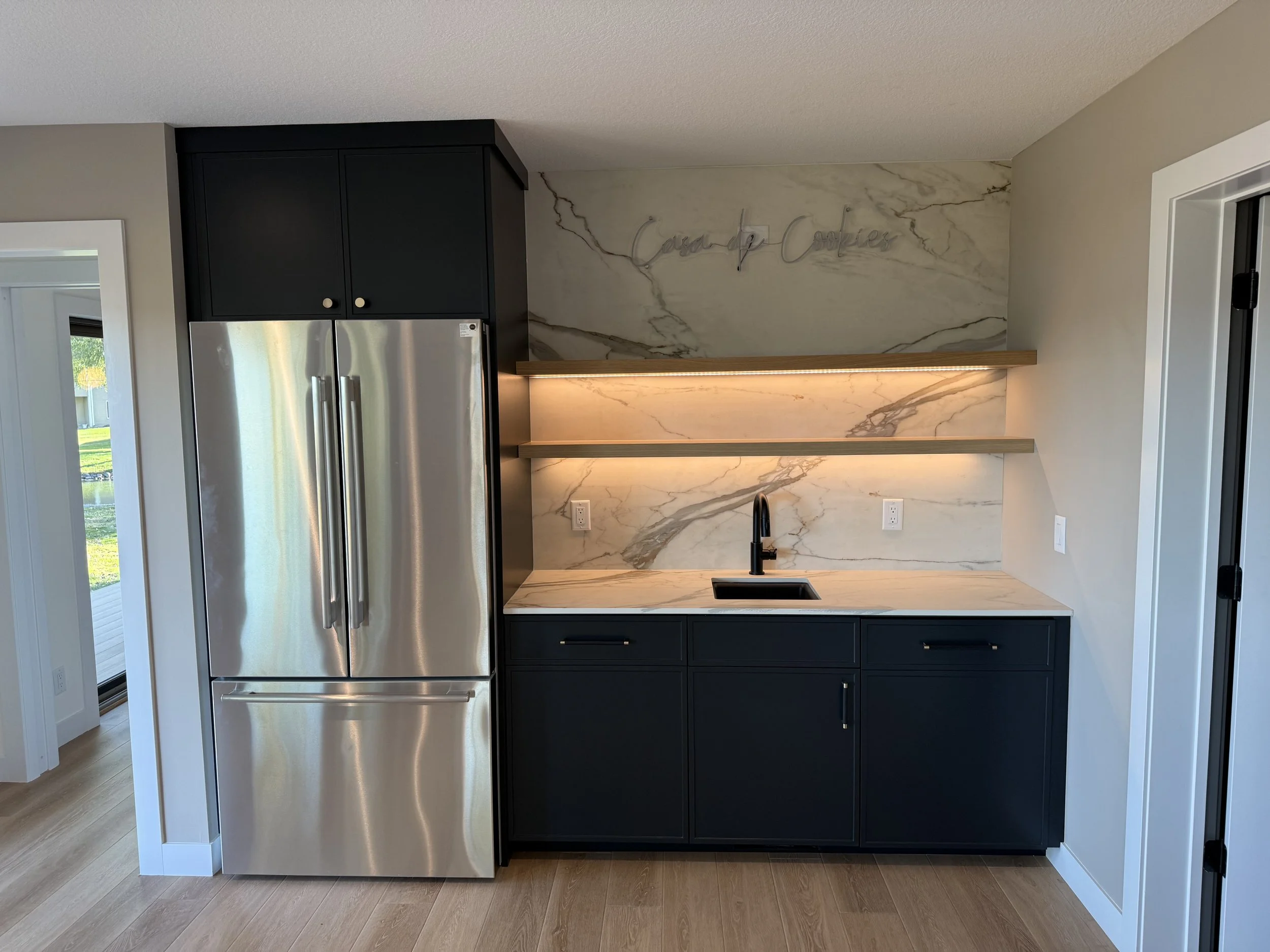 Modern kitchen with black cabinets, stainless steel refrigerator, marble backsplash, open shelves with lighting, and a small sink.