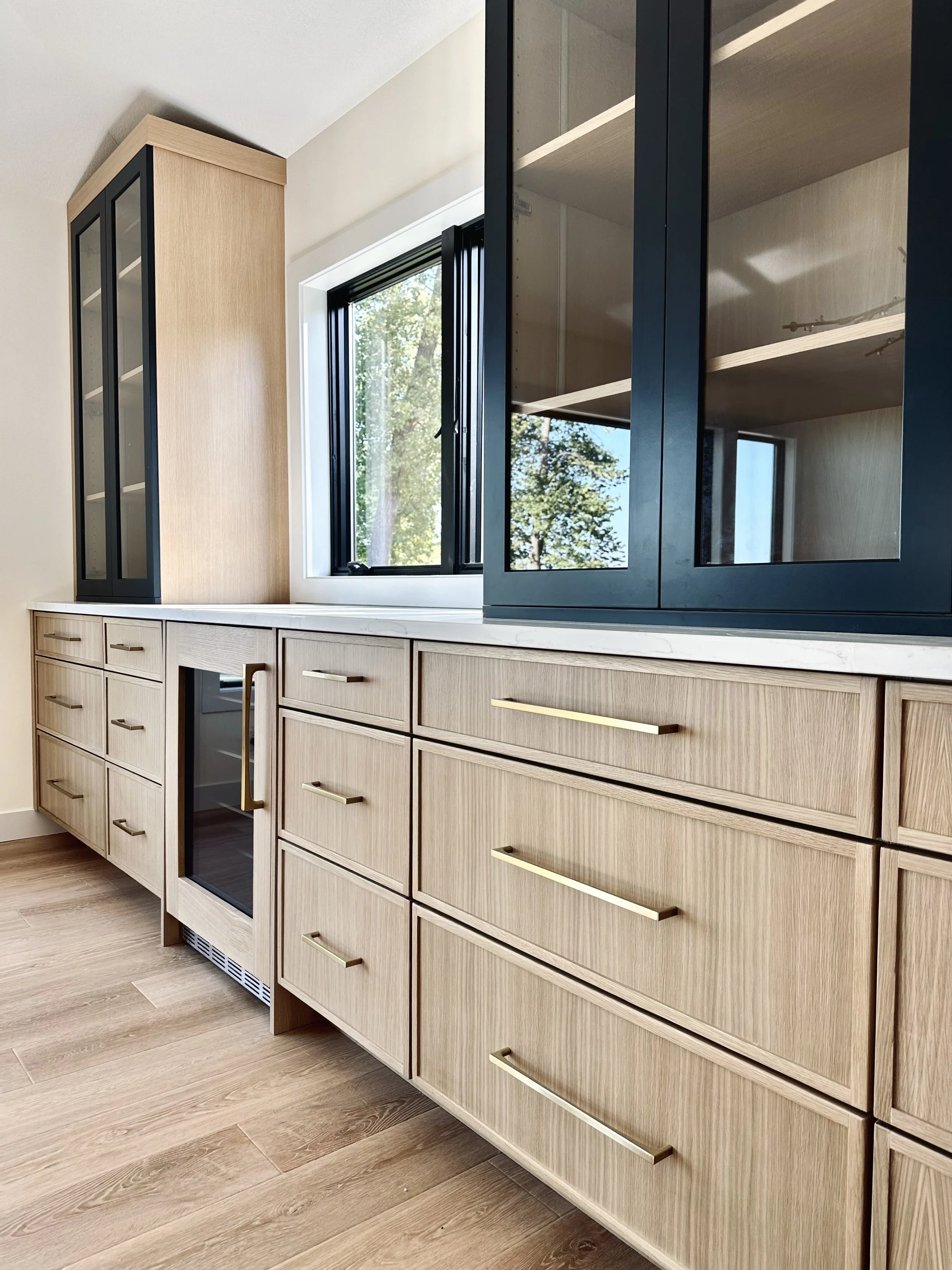 Modern kitchen with wooden cabinets, black framed glass doors, and an open window showing trees outside.