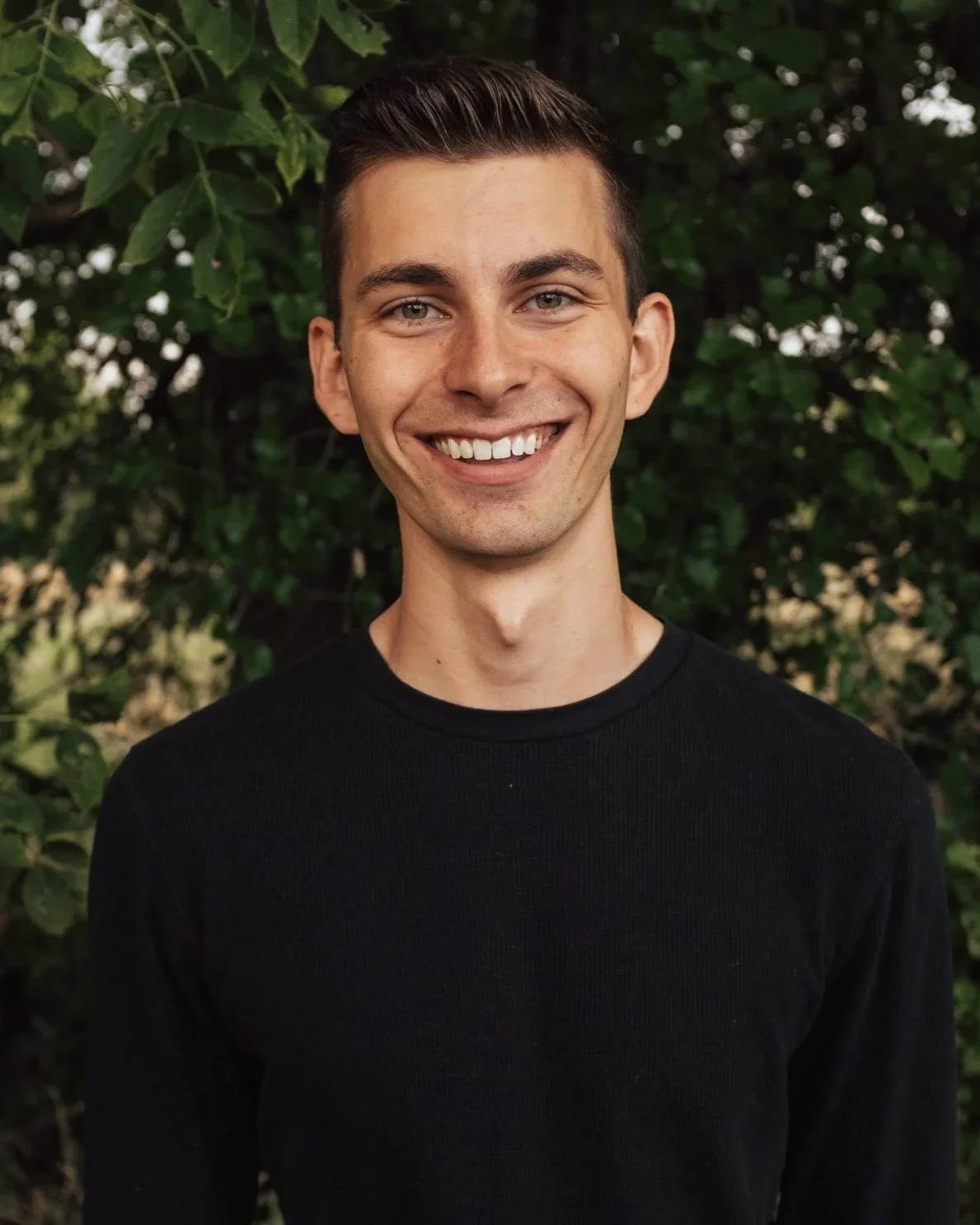 Smiling young man wearing a black shirt outdoors with green foliage background.
