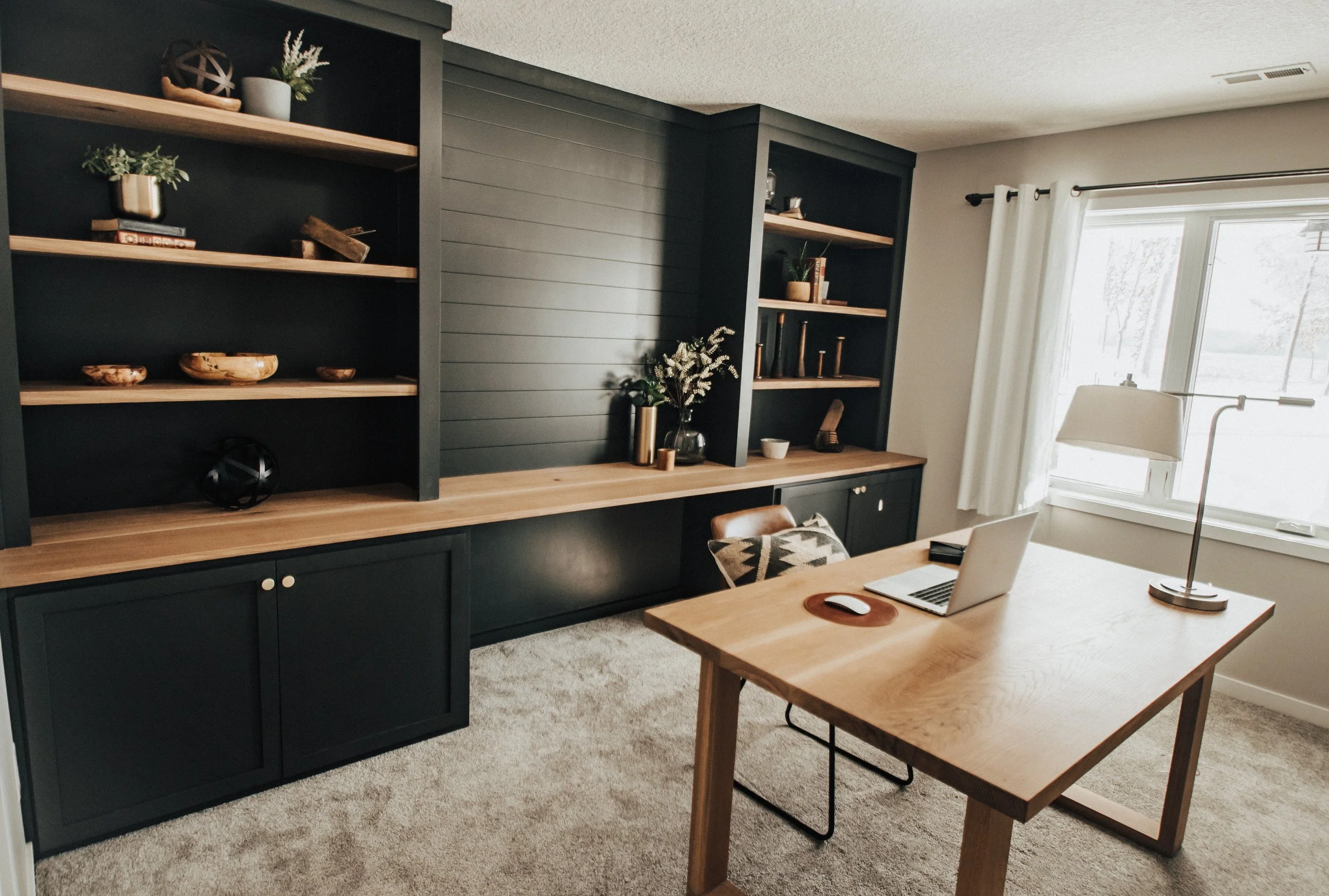 A home office with a wooden desk, a laptop, and a table lamp. Dark built-in bookshelves with decor and books, a large window with white curtains, and beige carpet flooring.