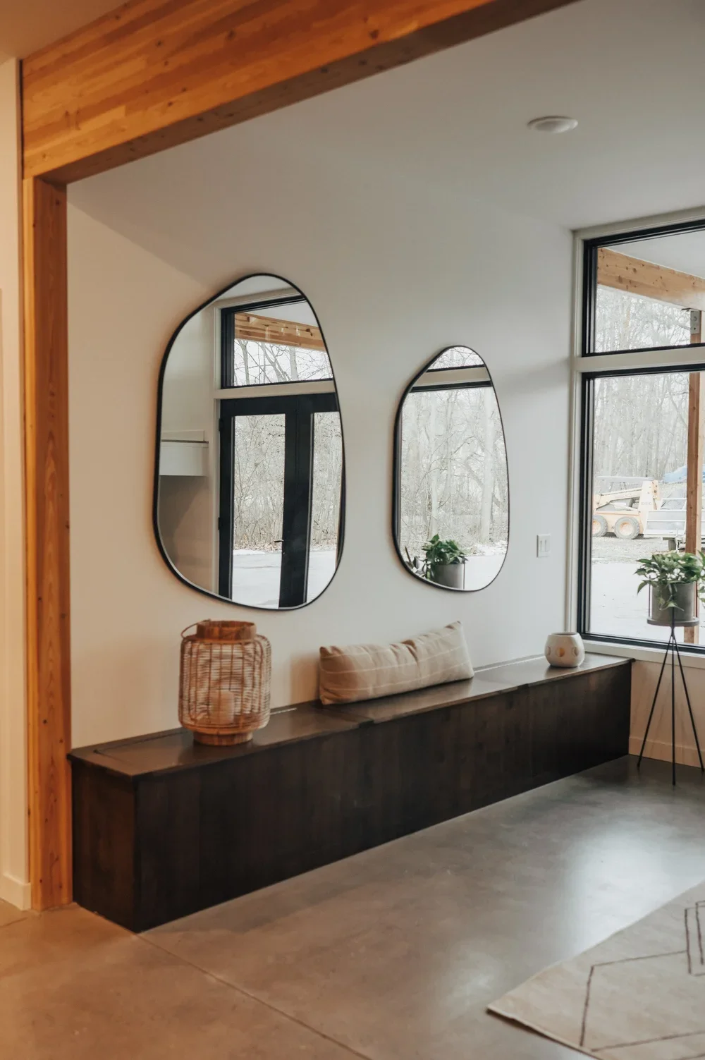 Interior of a modern room with two irregularly shaped mirrors on a white wall, a long dark wooden bench with a cushion, wicker and ceramic decorative items, and large windows with potted plants outside, showcasing a natural outdoor scene.