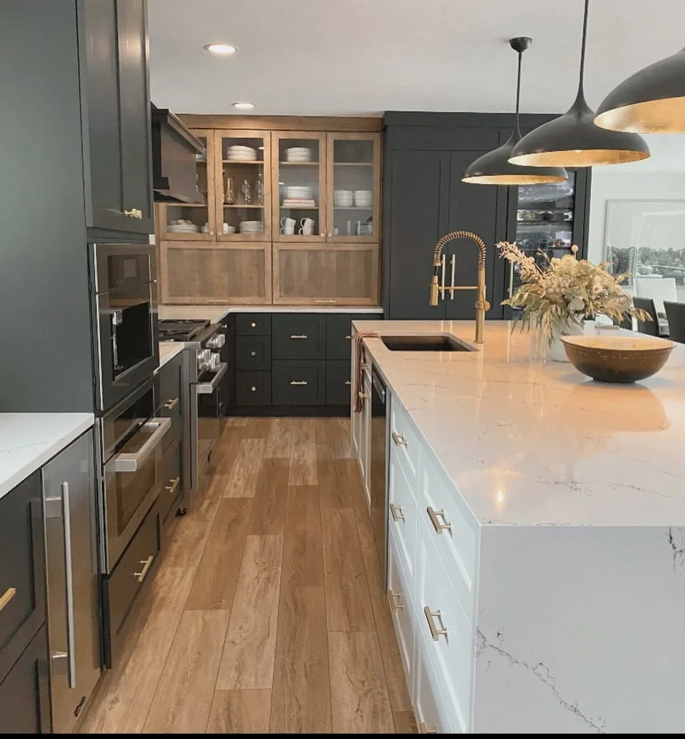 Modern kitchen with dark cabinets, light marble island, wooden flooring, open shelving with dishes, and gold accents.