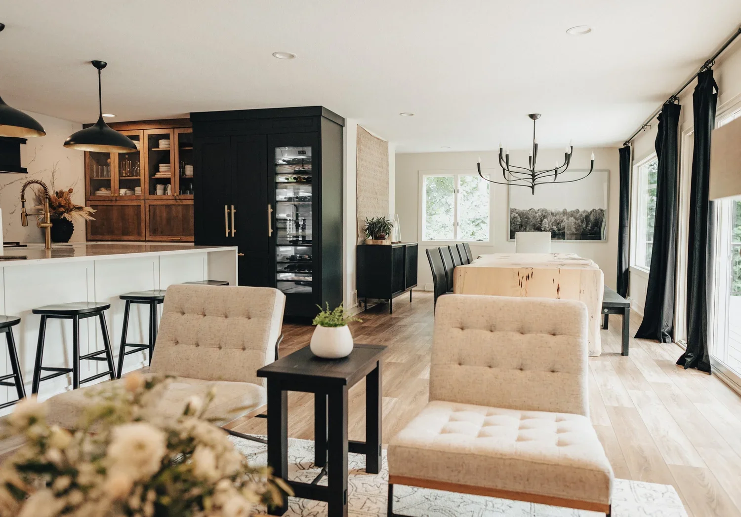Open-concept living area with dining table, beige chairs, black chairs, black cabinet, and kitchen island with barstools, hardwood flooring, and large windows with black curtains.