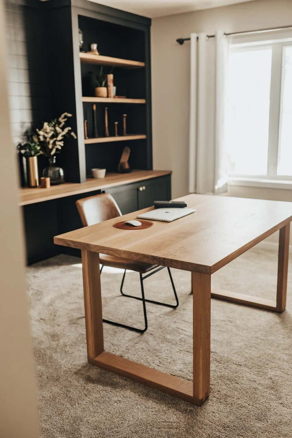 A modern home office with a wooden desk, brown leather chair, a laptop, mouse, remote, and phone on the desk. There are white curtains covering a large window, and a black shelving unit with decorative items, books, and candles behind the desk. The room has beige carpet and white walls.
