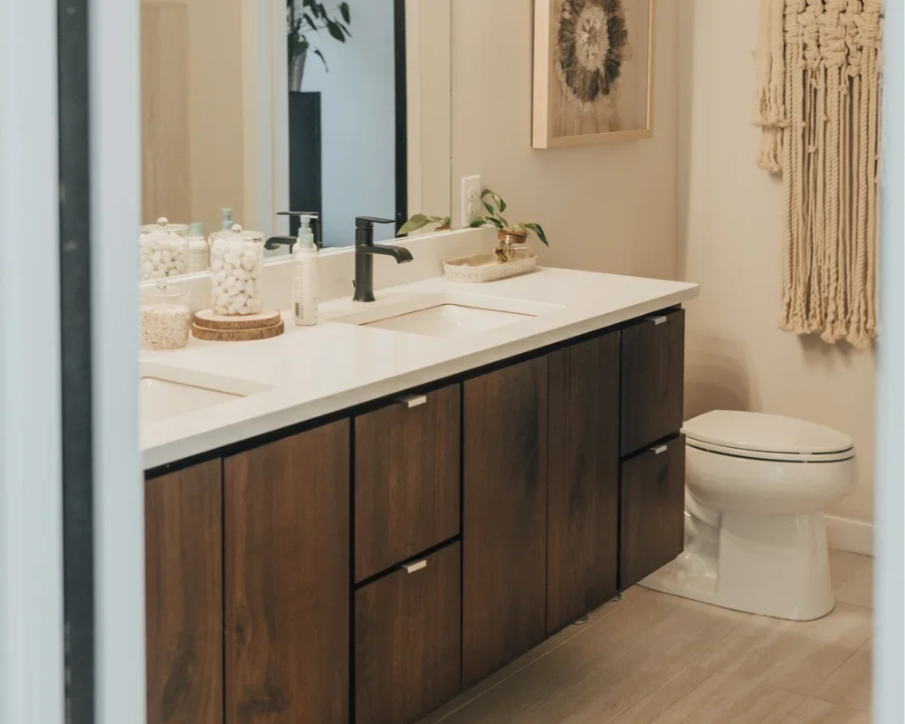 Modern bathroom with double vanity featuring dark wood cabinets, white countertop, and black faucet, mirror, soap dispenser, jars, framed wall art, macramé wall hanging, and toilet.