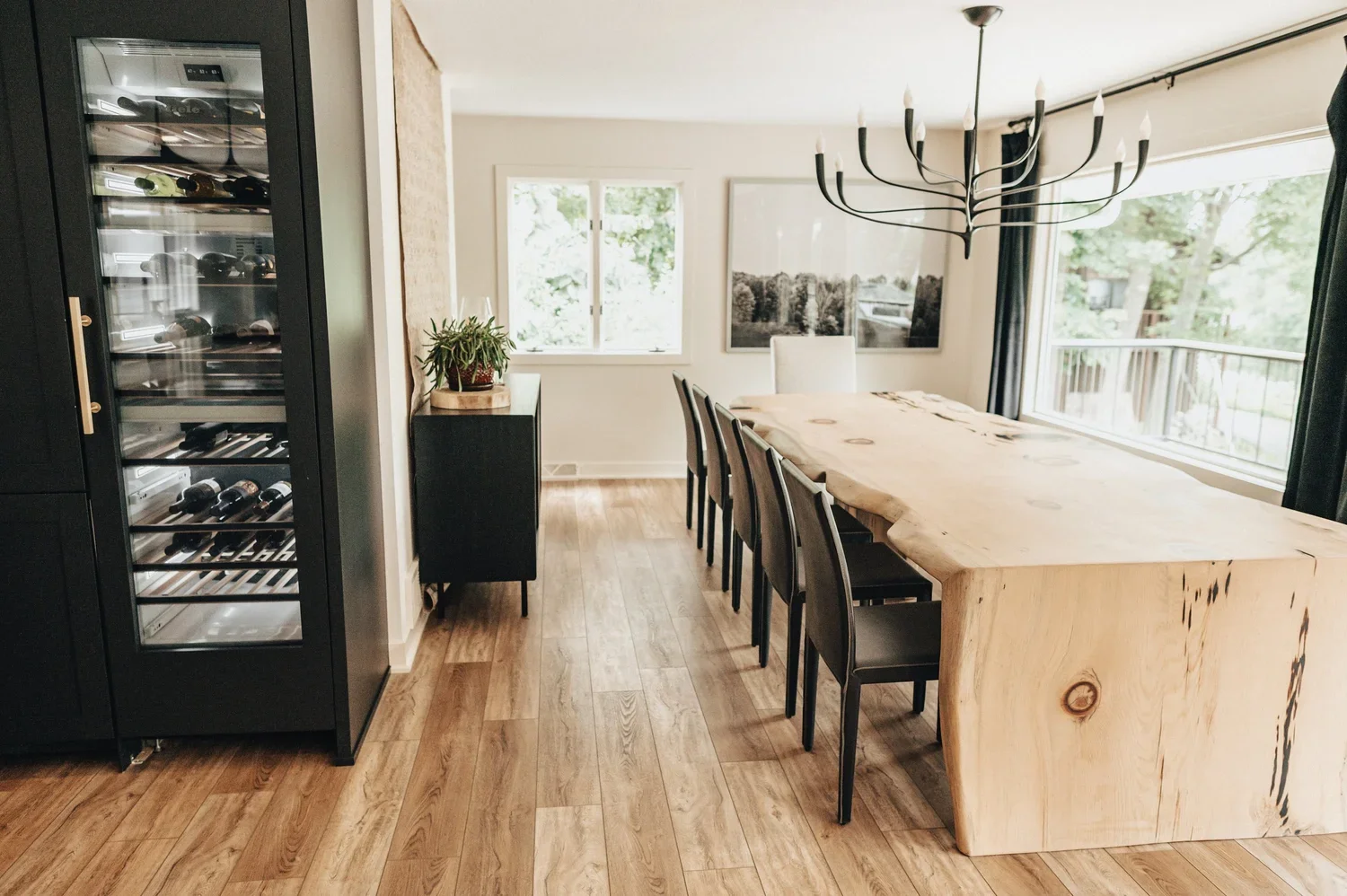 A dining room with a large wooden table, six black chairs, a black chandelier, a window with outside greenery, a black cabinet with a coffee machine, and a black sideboard with a plant on top.
