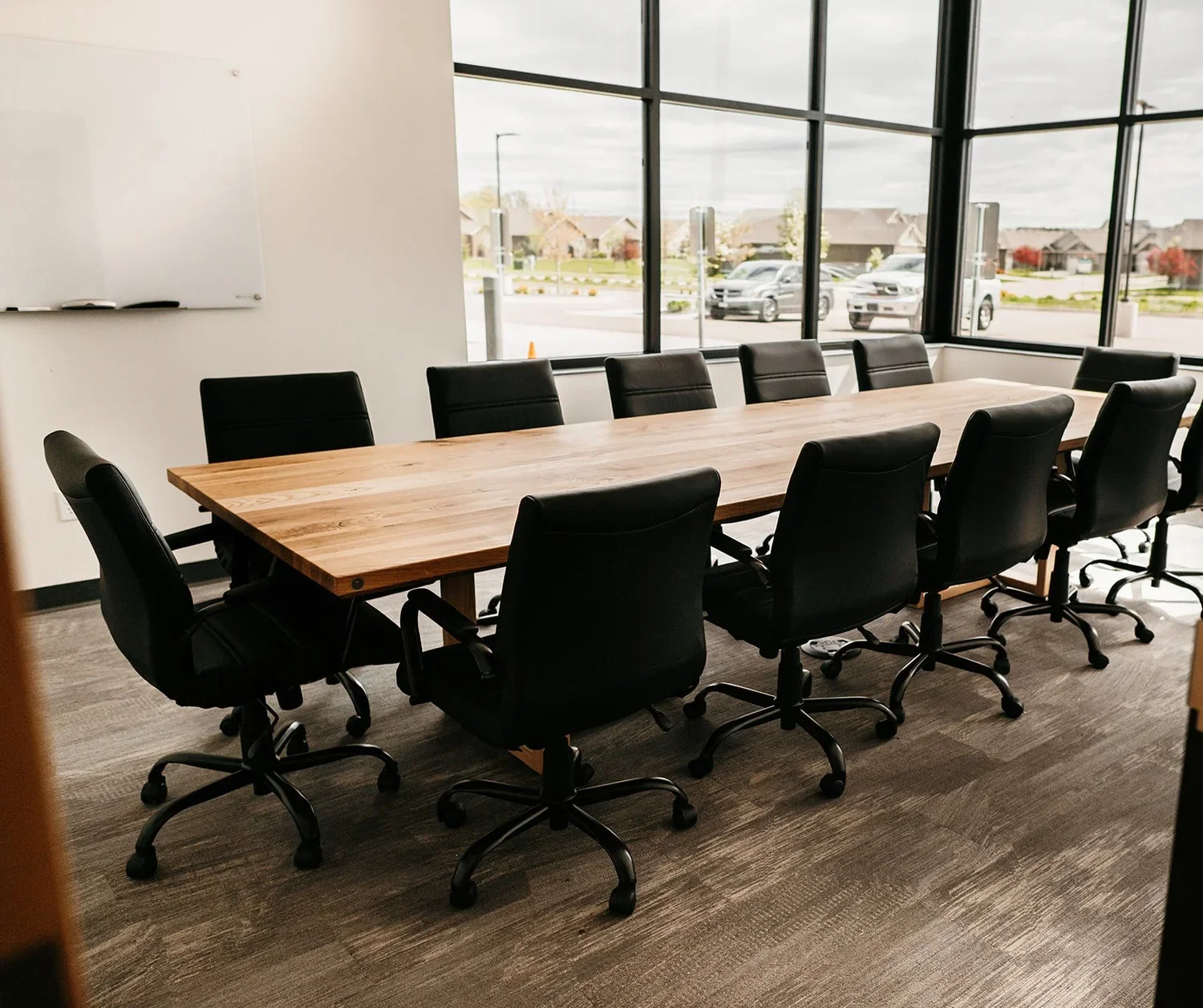Conference room with a large wooden table and twelve black office chairs, with large windows showing cars outside.