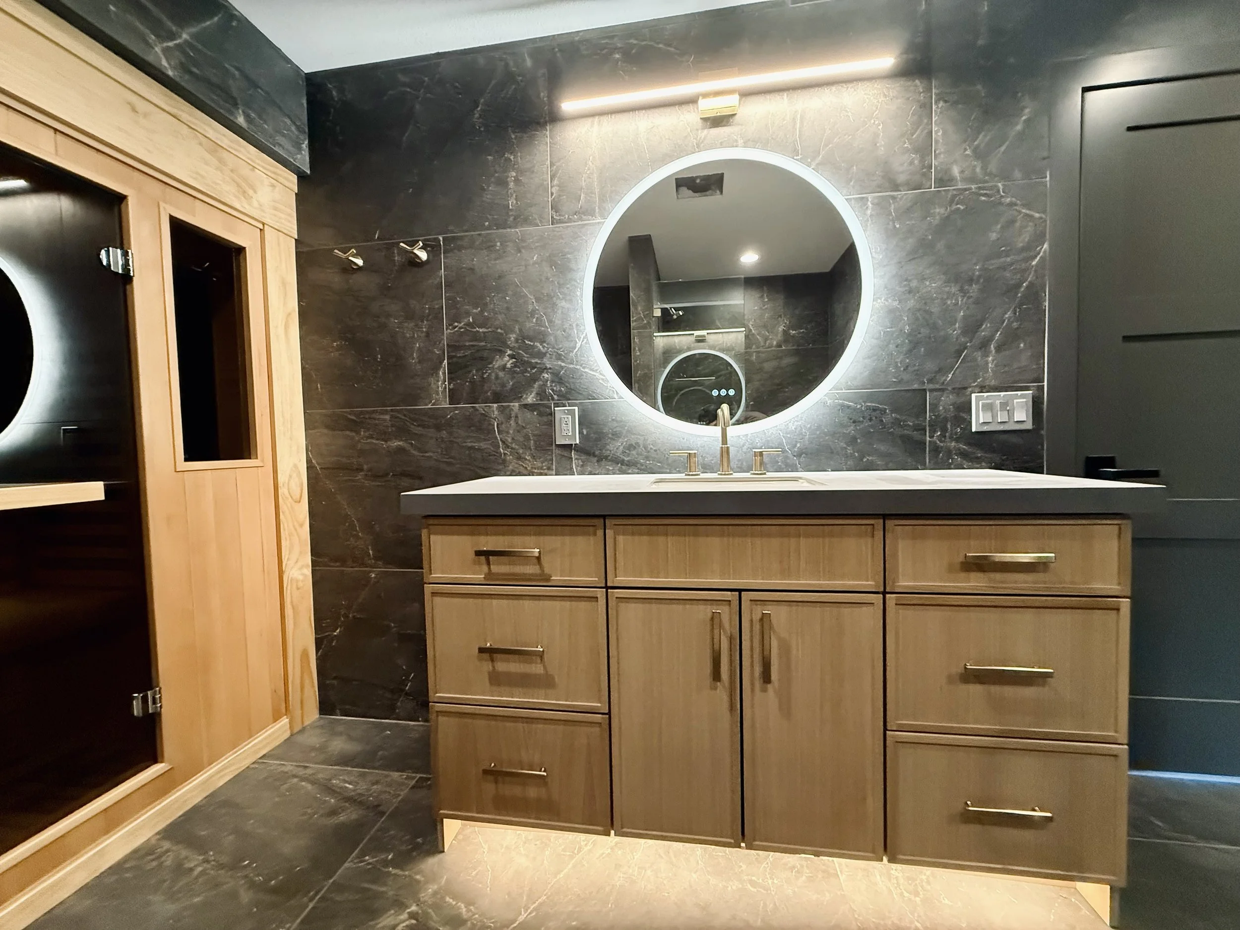 Modern bathroom vanity with a round backlit mirror, black marble wall, wooden cabinets with gold handles, gray countertop, and a European-style sauna to the left.