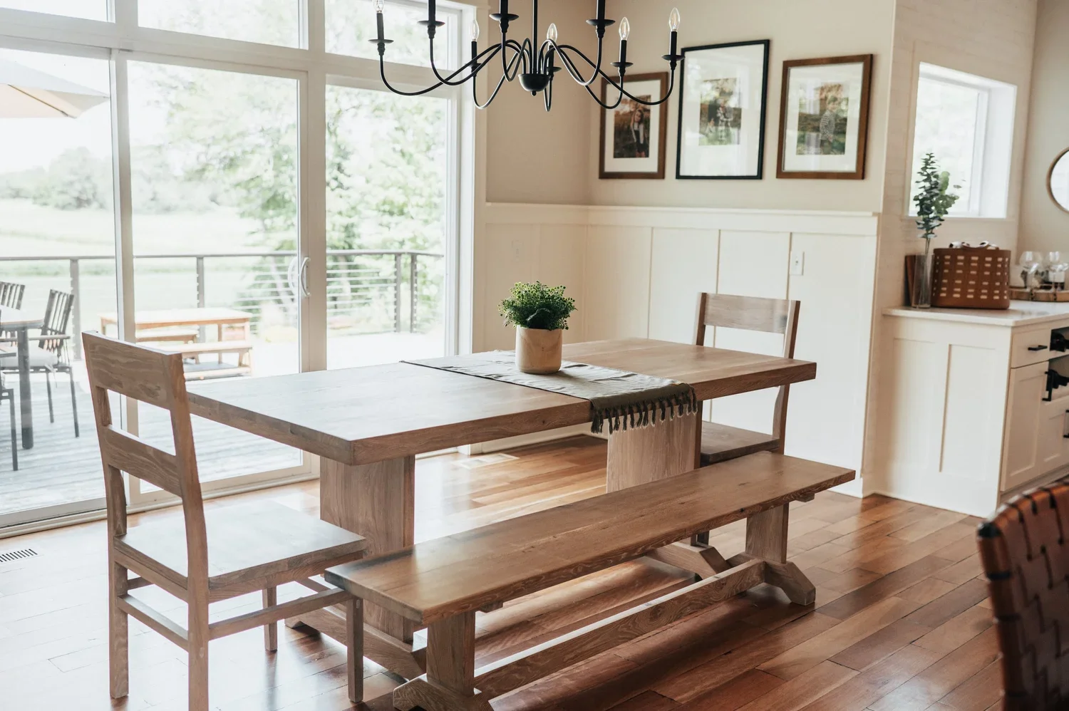 Dining area with wooden table, bench, and chairs near large sliding glass doors leading to a deck with outdoor furniture. The room has framed photos on the wall, a chandelier hanging over the table, and a small plant in a pot as a centerpiece.