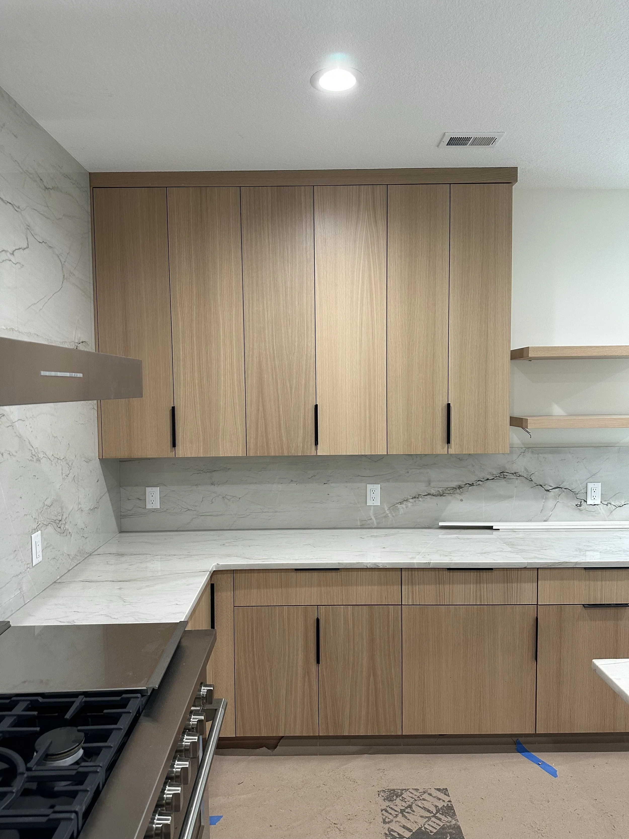 Kitchen with wooden cabinets, marble countertops, and a stainless steel stove, with outlets on the backsplash and open shelves on the right.