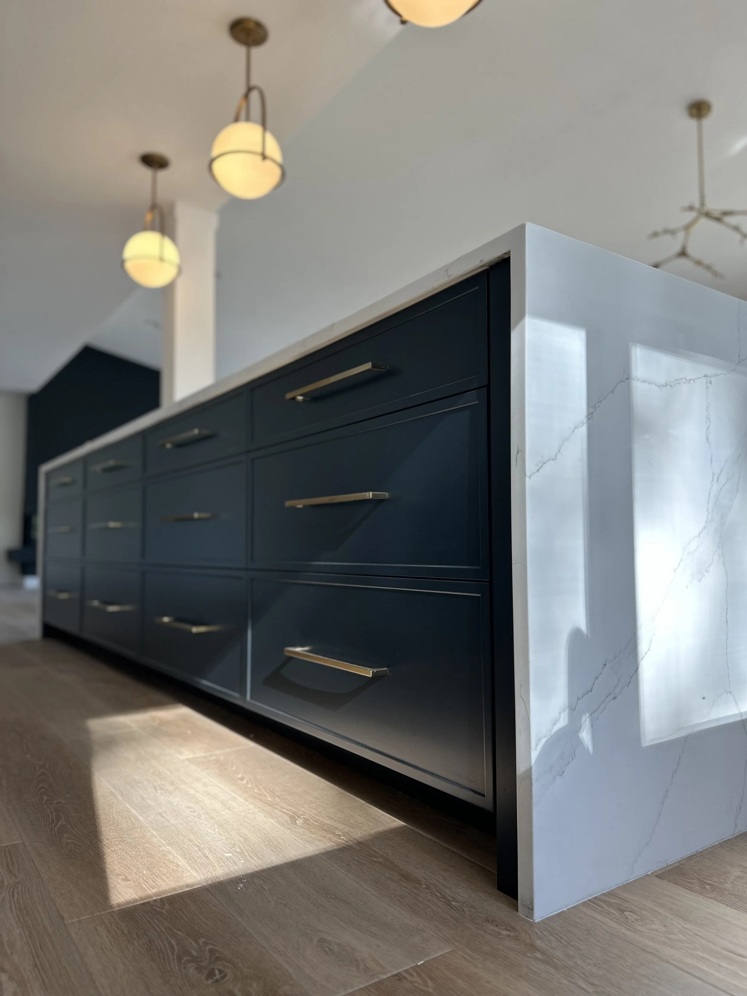 Close-up of a modern kitchen island with dark blue cabinets and gold handles, topped with white marble countertop, against a background of a room with pendant lights and a ceiling fixture.