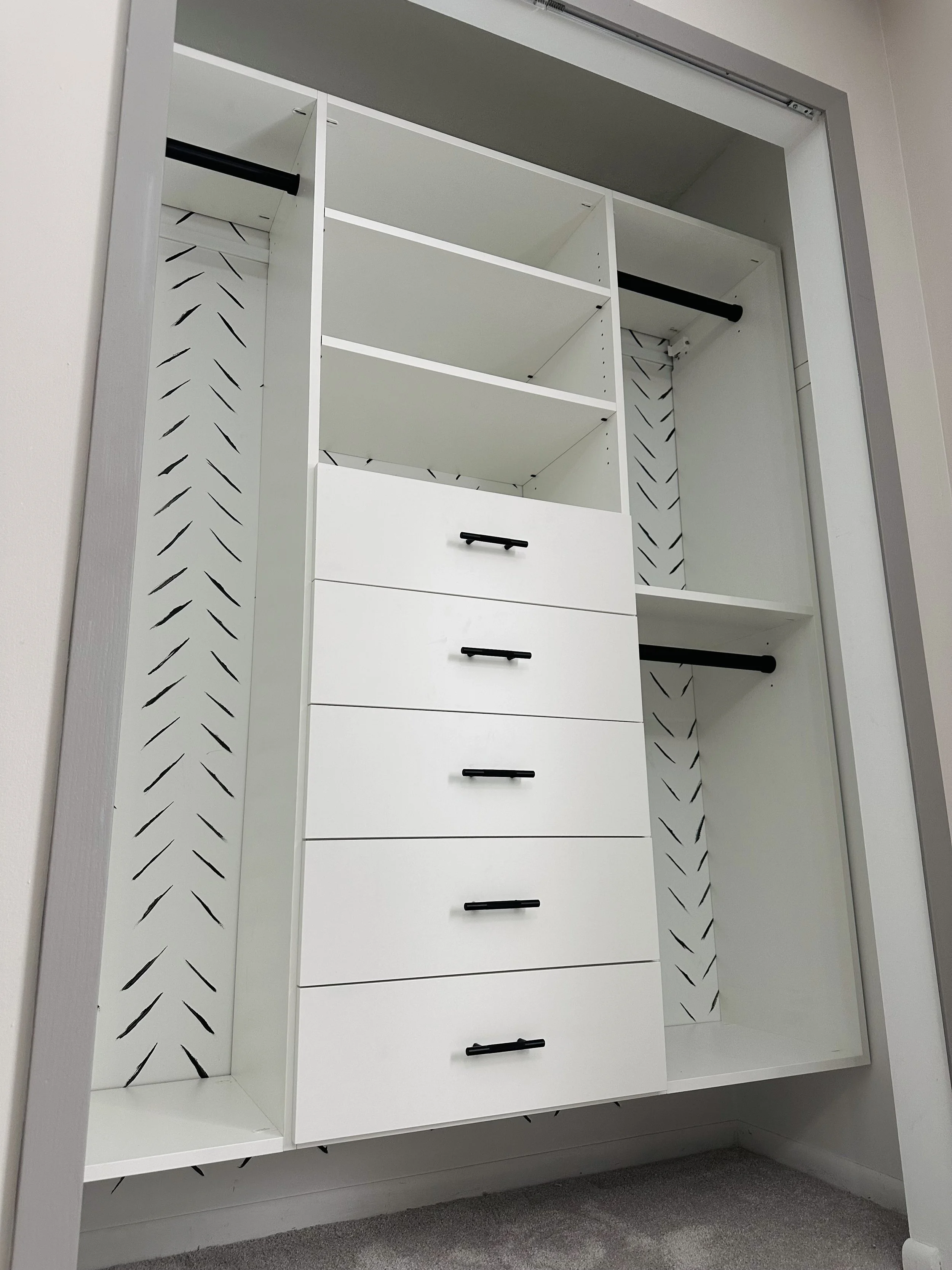 Empty white closet with multiple shelves and five drawers, black handles, with black rods for hanging clothes.