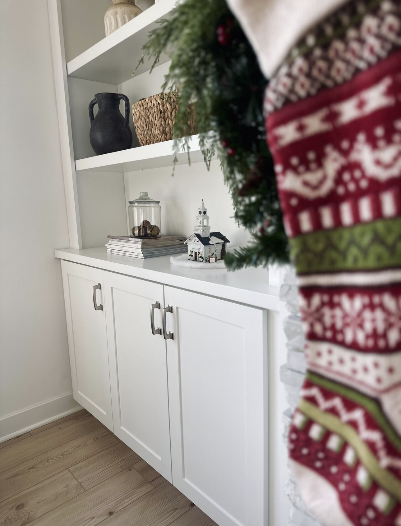 Part of a white cabinet with a holiday decoration partly visible in the foreground; background shows white shelves with vases, a jar of nuts, and a small decorative church.