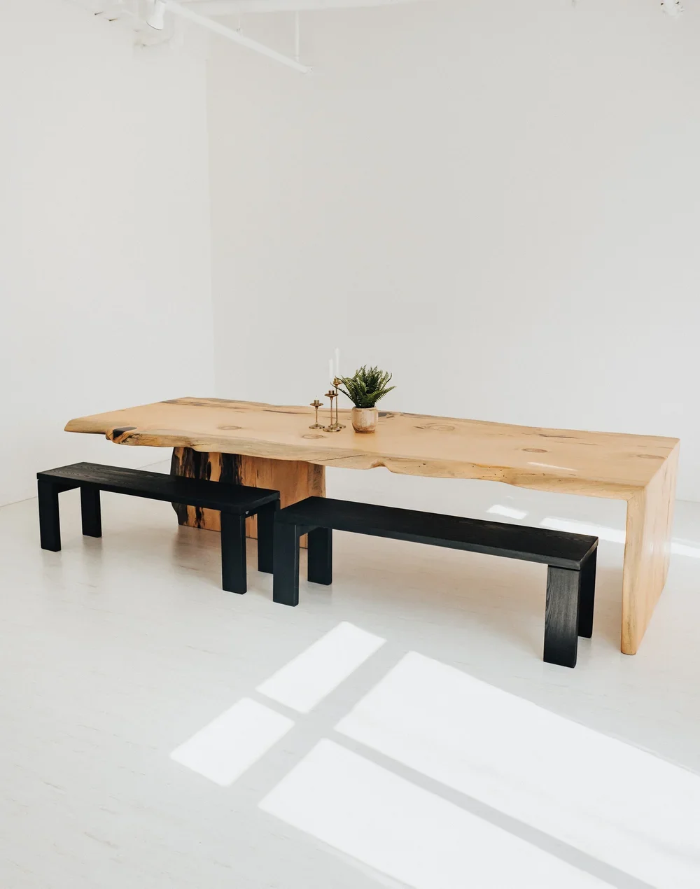 Minimalist room with a natural wood dining table, two black benches, a potted plant, and candlesticks, illuminated by sunlight casting shadows on a white floor.