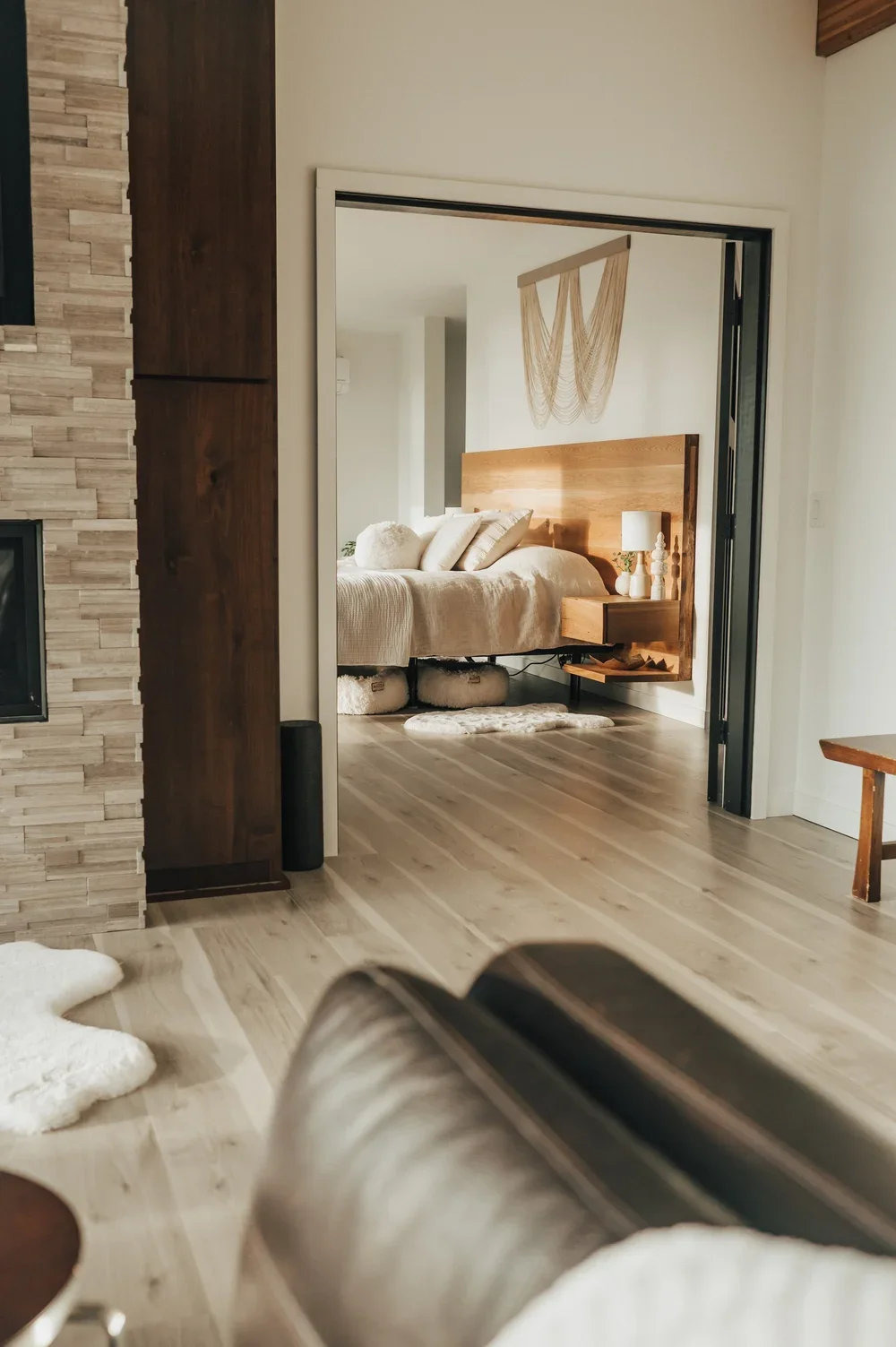 A bedroom with a wooden headboard, beige bedding, pillows, a nightstand with a lamp, a wood wall decor piece, and a cowhide rug. The room is visible through a doorway from a living area with a partial view of a fireplace and floor.