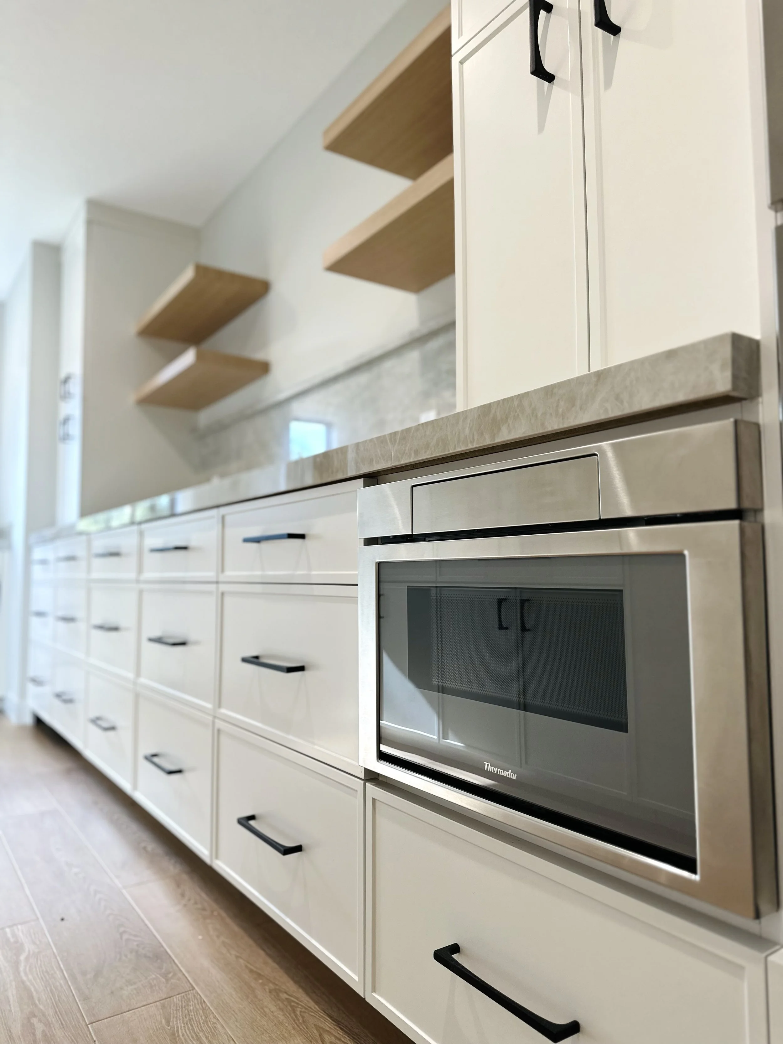 Modern kitchen with white cabinets, black handles, built-in microwave, and open wooden shelves.