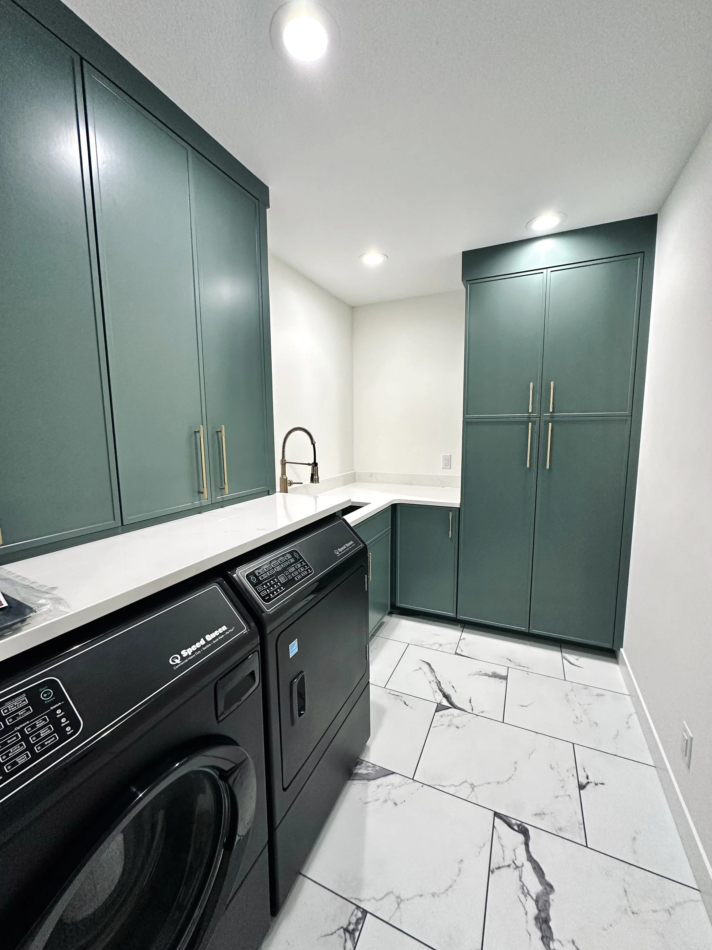 Modern laundry room with green cabinets, white countertops, a black washing machine and dryer, marble floor tiles, and white walls.