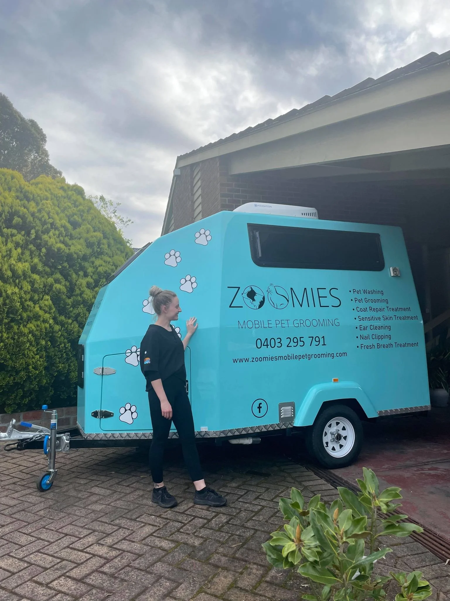 A woman standing next to a blue pet grooming trailer with paw prints and company information printed on it, parked on a brick driveway.