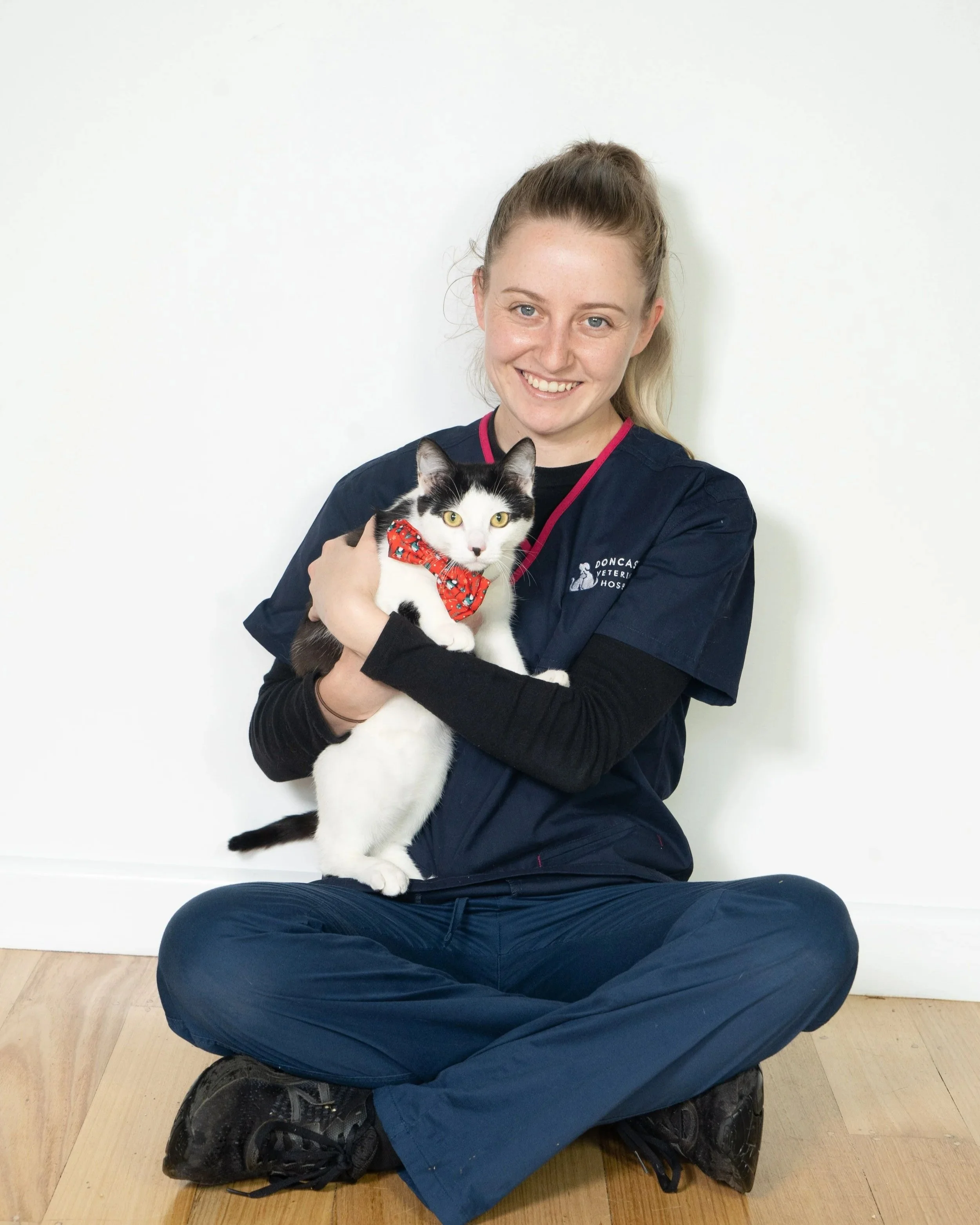 A woman in veterinary scrubs sitting cross-legged on the floor, holding a black and white cat with a red bow tie. She is smiling at the camera against a plain white wall.