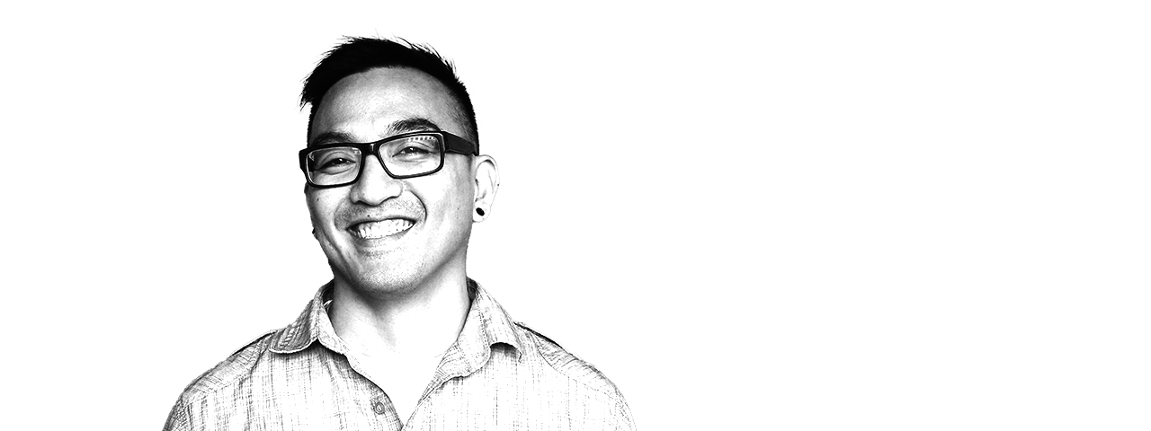 A smiling man with glasses and a short hairstyle, wearing a button-up shirt, standing against a background with black and gray horizontal stripes.