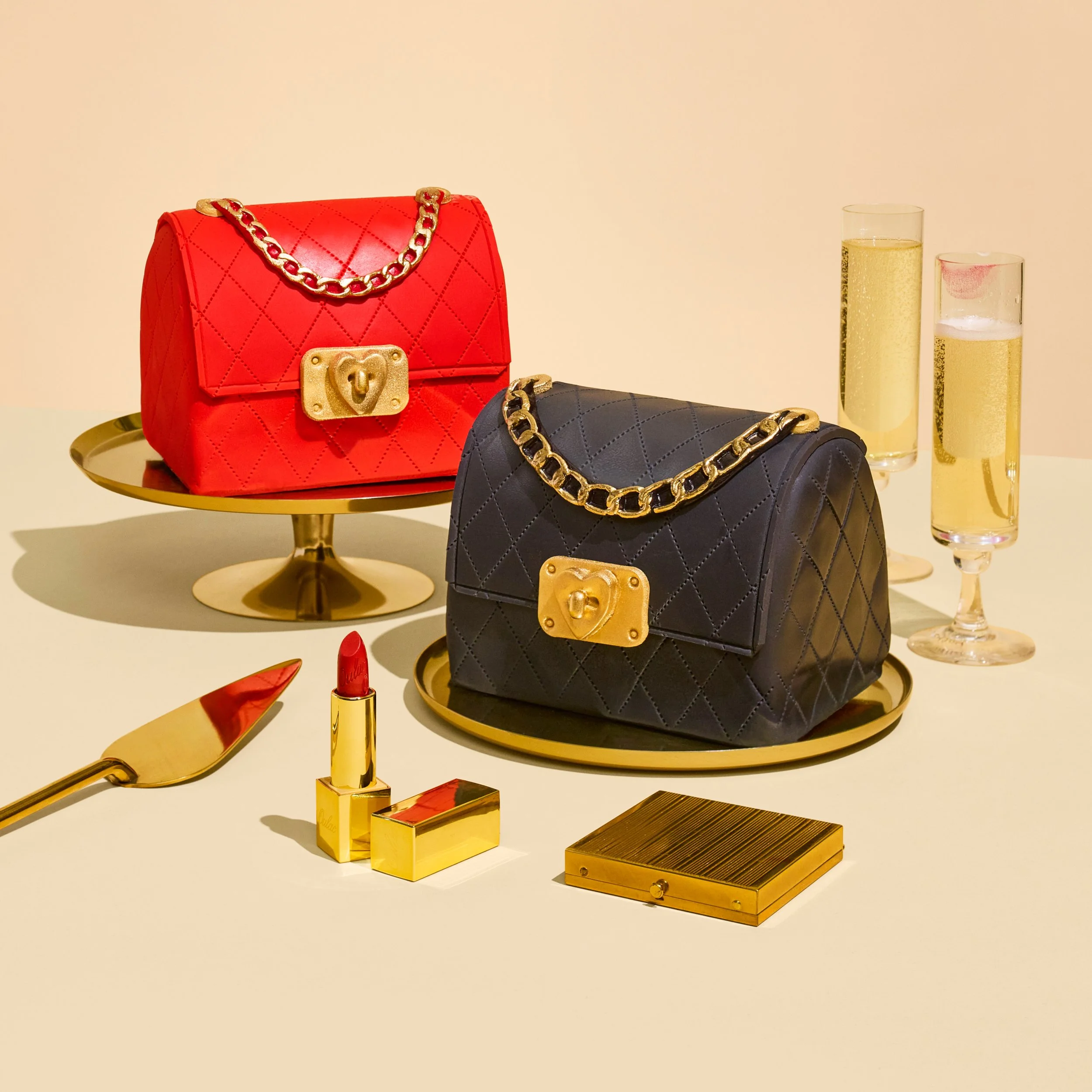 Two handbags, one red and one black, with gold chains and clasps, displayed on gold trays. The red purse is on a raised stand, while the black purse is directly on the surface. Nearby are two glasses of champagne, a gold lipstick, a gold lipstick cas