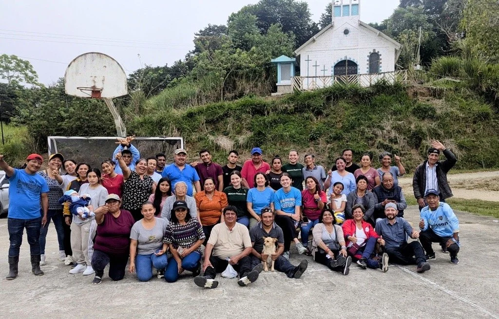 Group of people gathered on a basketball court outdoors, with a small dog in front, and a church building on a hill in the background.