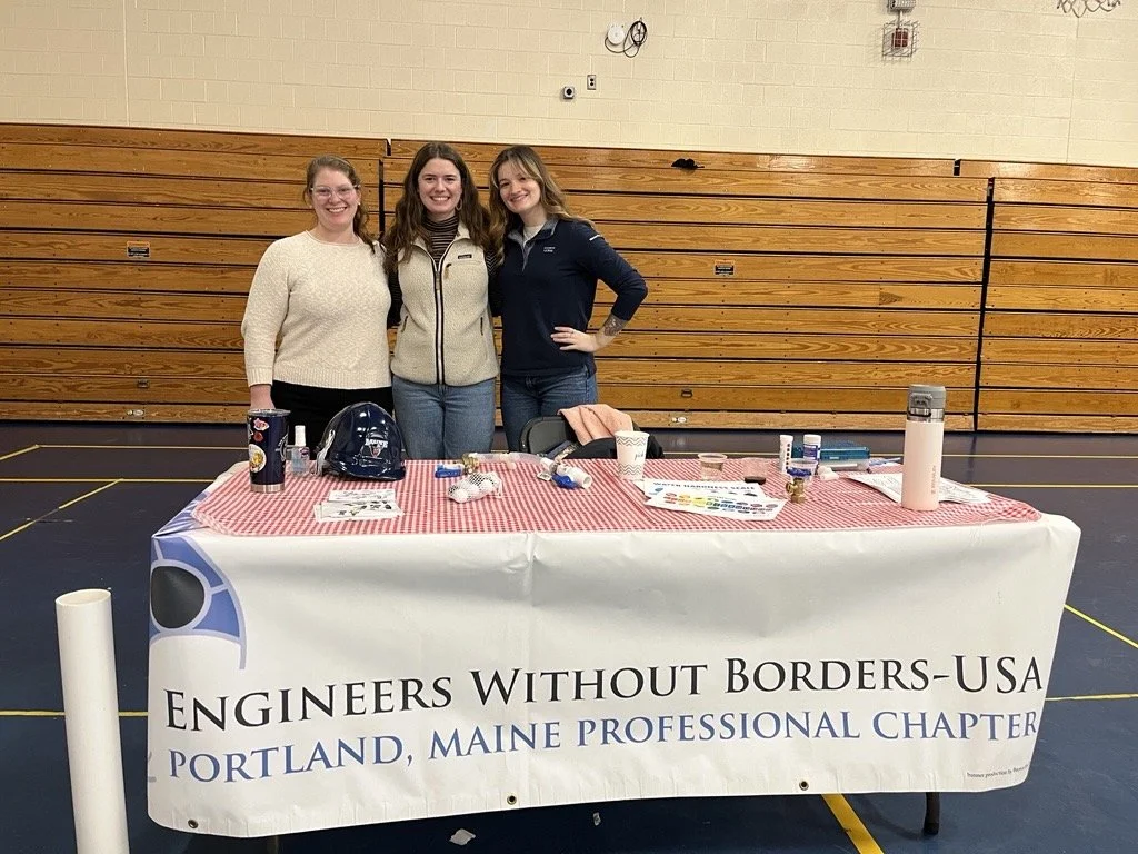 Three people standing behind a table with display boards and framed photographs at an event. The table is covered with a banner that reads 'Engineers Without Borders'.