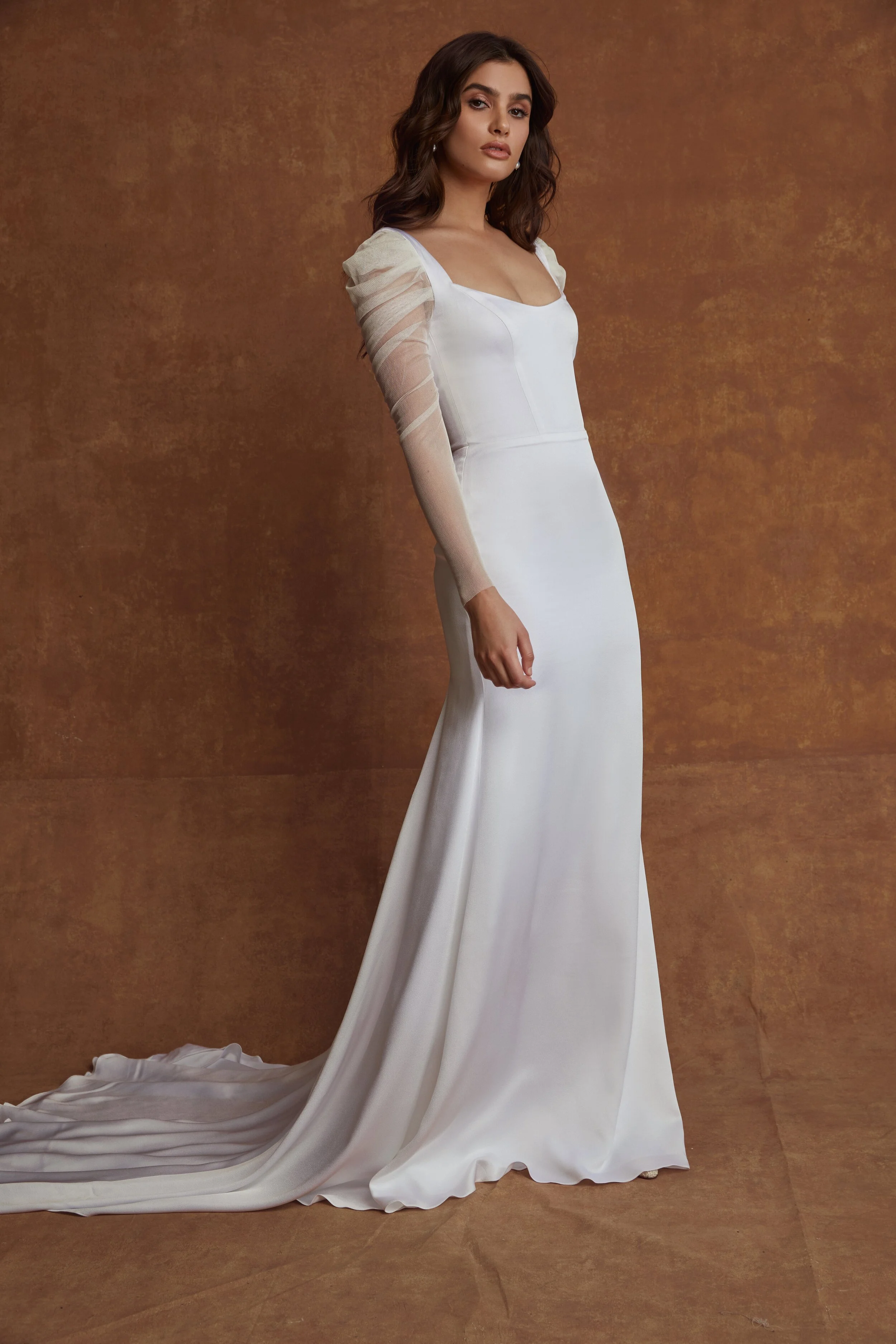 A young woman in a white wedding gown with sheer puffed sleeves and a long train, standing against a textured brown background.