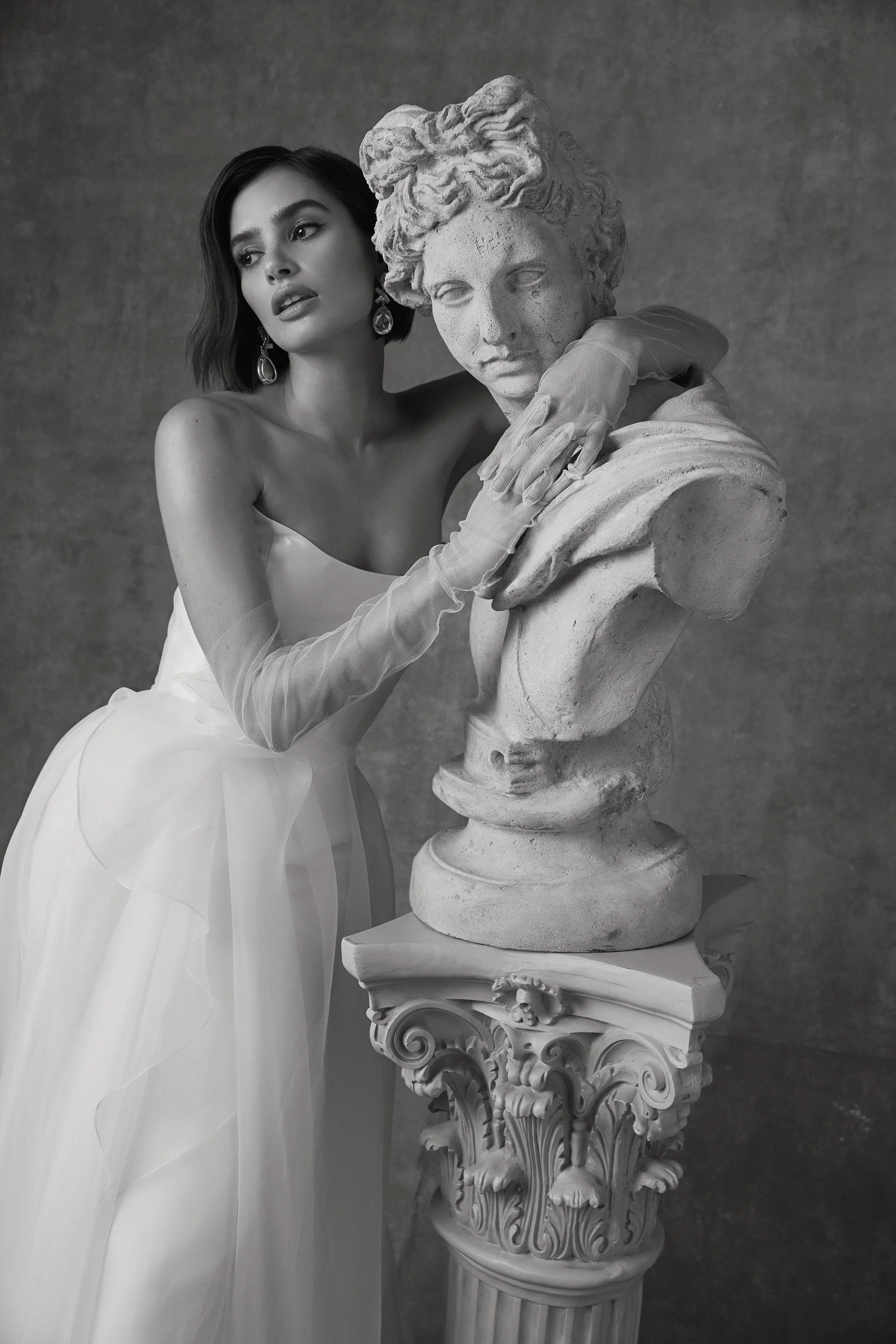 A woman in a white dress and long gloves standing next to a classical marble bust sculpture of a woman, both with serious expressions, in a black-and-white photograph.