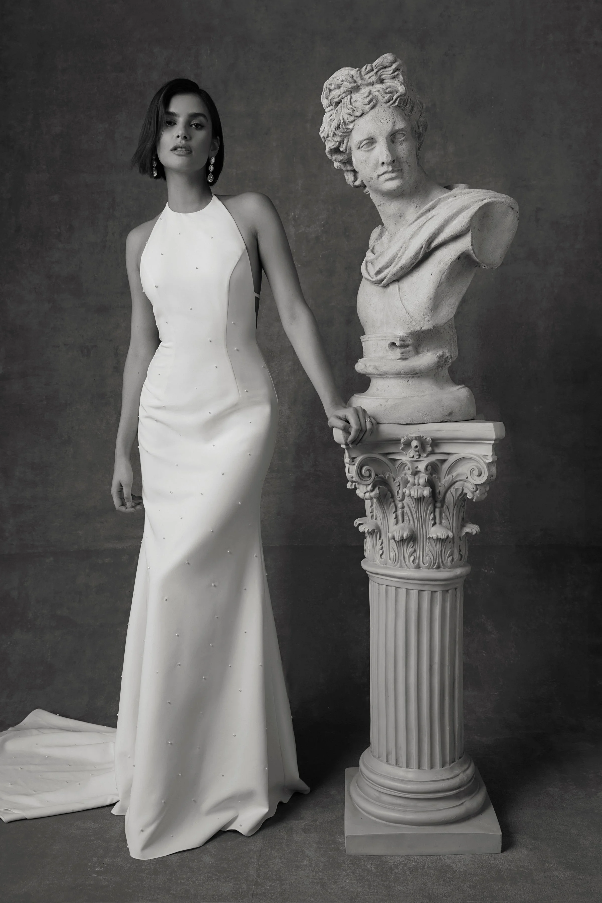 A woman in a white dress standing next to a classical sculpture bust on a decorative pedestal.