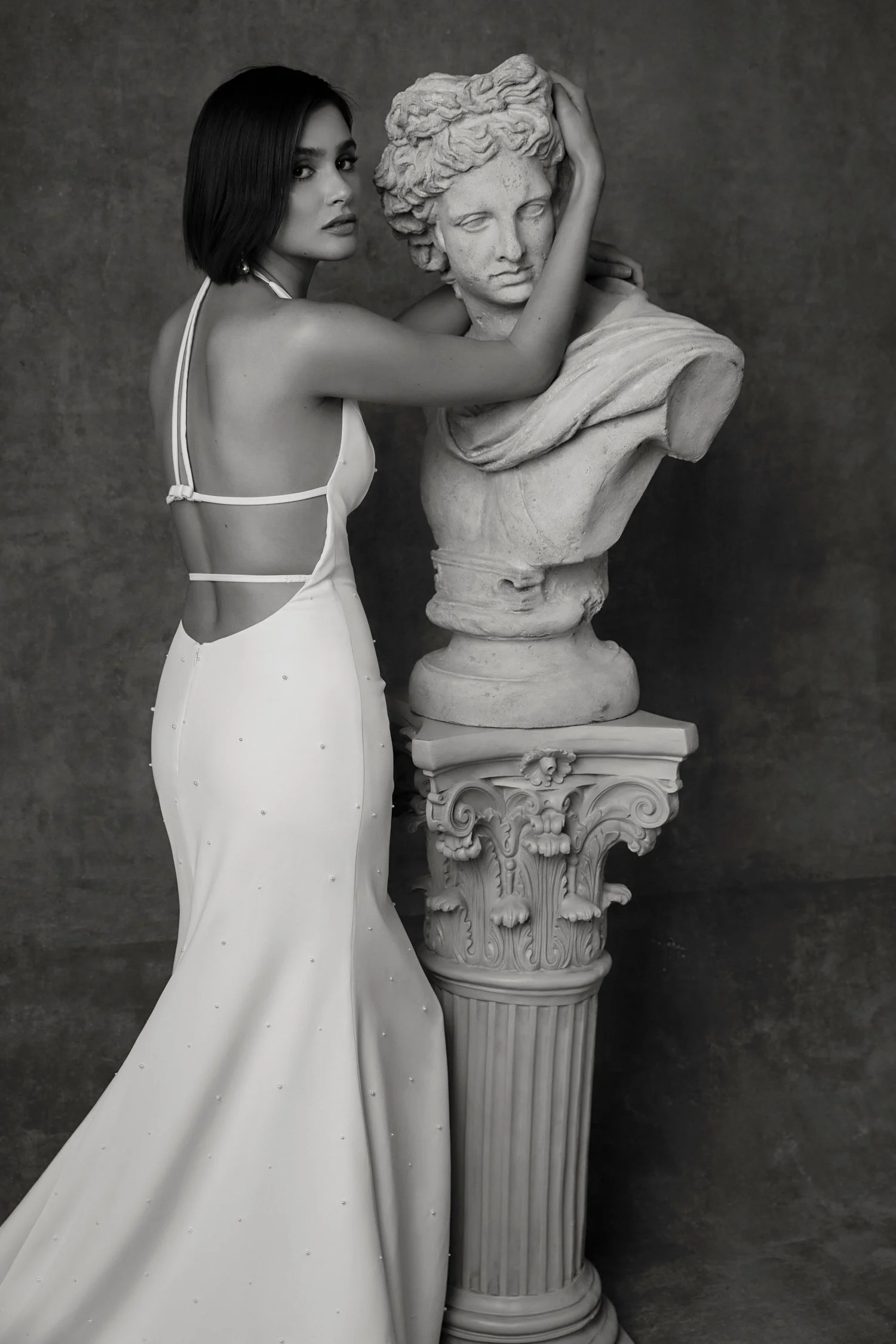 A woman in a white gown with open back and thin straps poses with a classical bust sculpture on a pedestal.