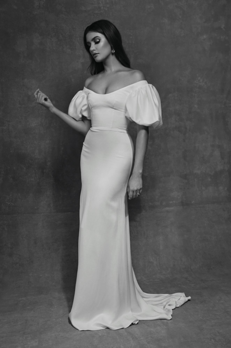A woman in an off-the-shoulder, floor-length white gown with puffy sleeves, standing against a textured wall in black and white.