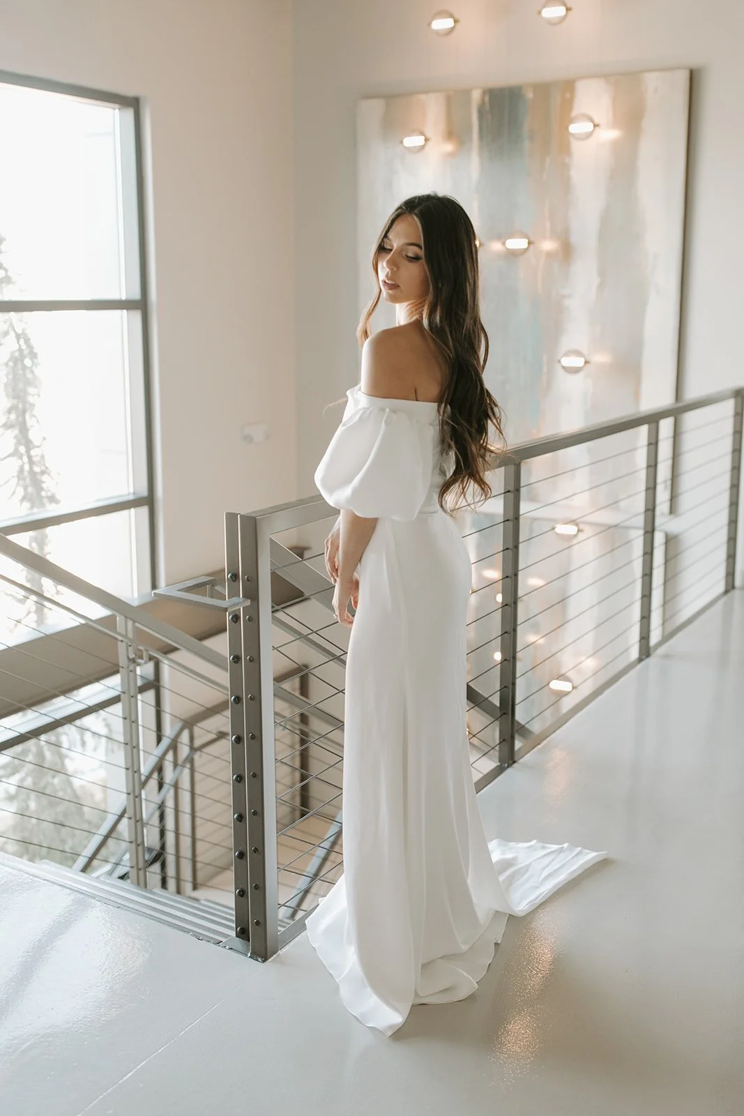 A woman in a white off-the-shoulder dress standing on a balcony with a modern interior background, large windows, and natural light.