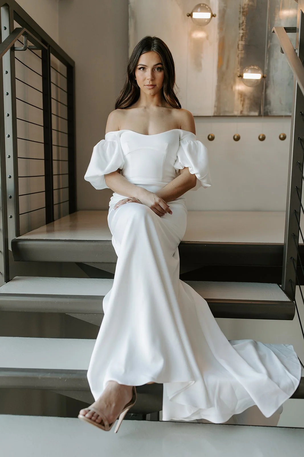A woman in a white off-shoulder gown sitting on a modern staircase in an indoor setting.