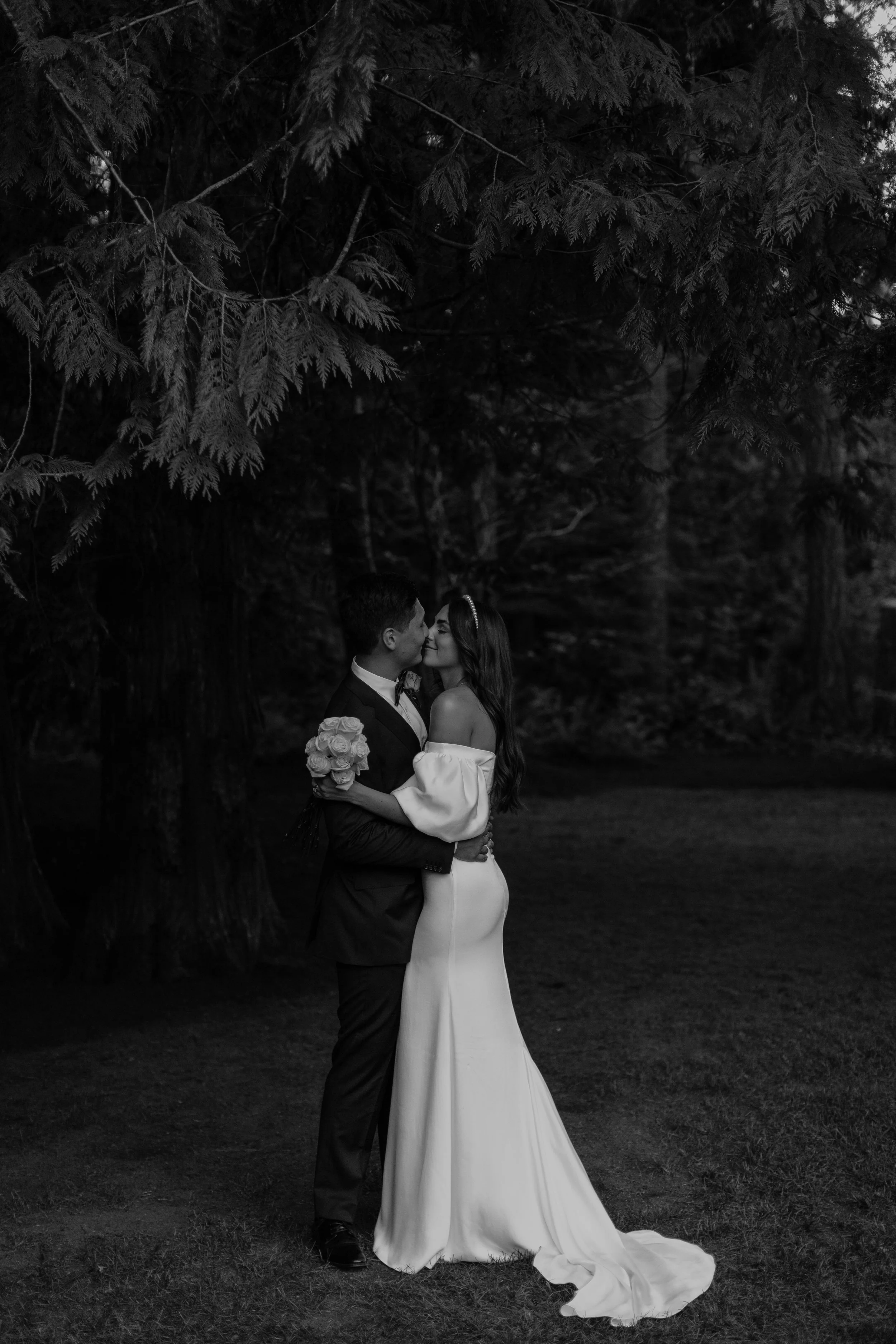 Black and white photo of a bride and groom embracing and kissing under a large tree in an outdoor setting, with grass on the ground.