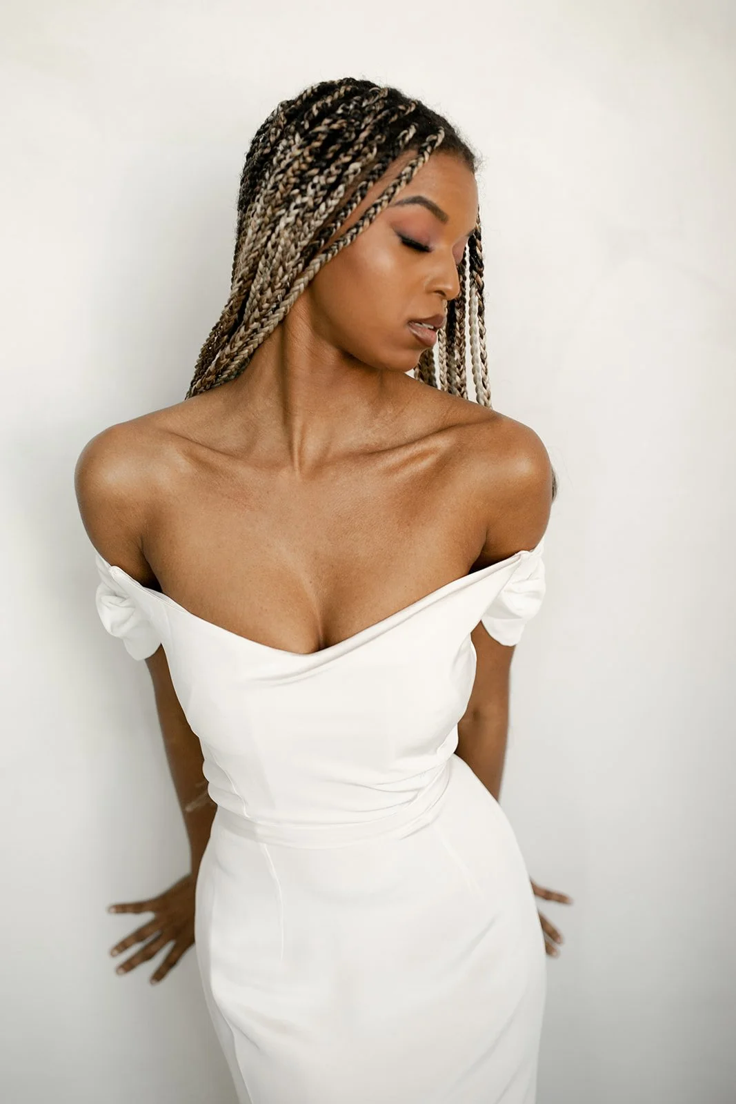 A woman with braided hair in a white off-shoulder dress, standing against a plain background.