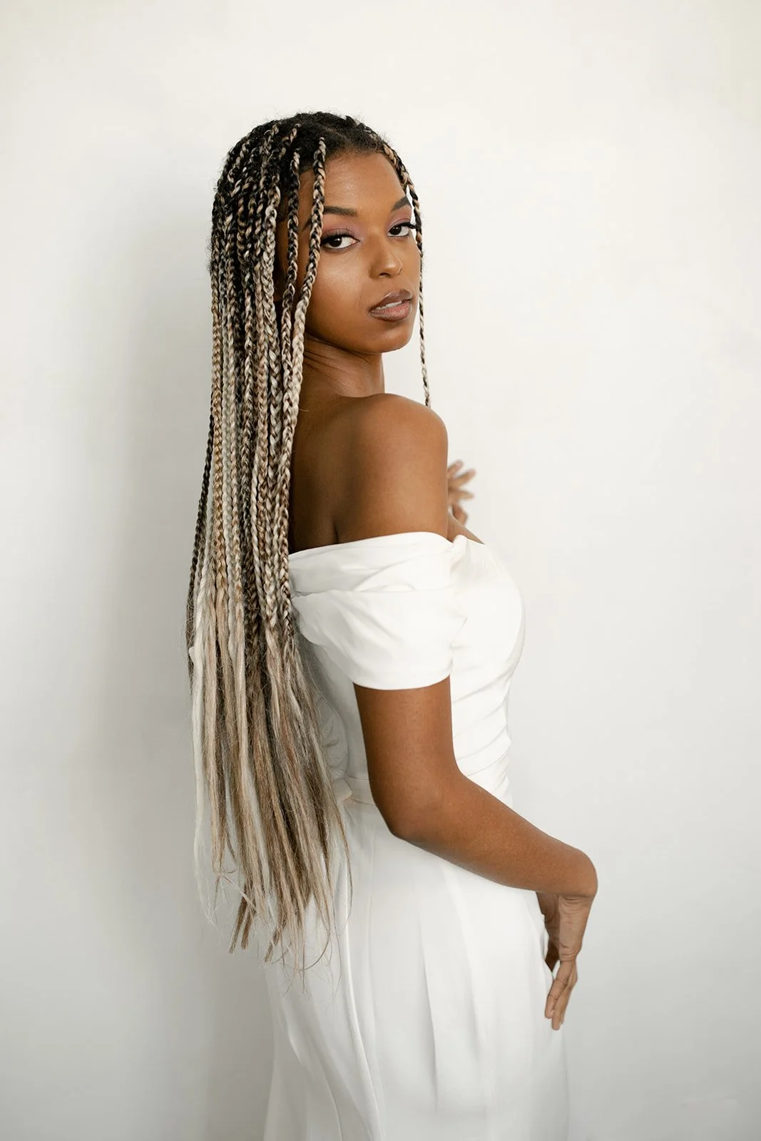 A woman with long braided hair, wearing a white off-shoulder dress, standing against a plain white background.